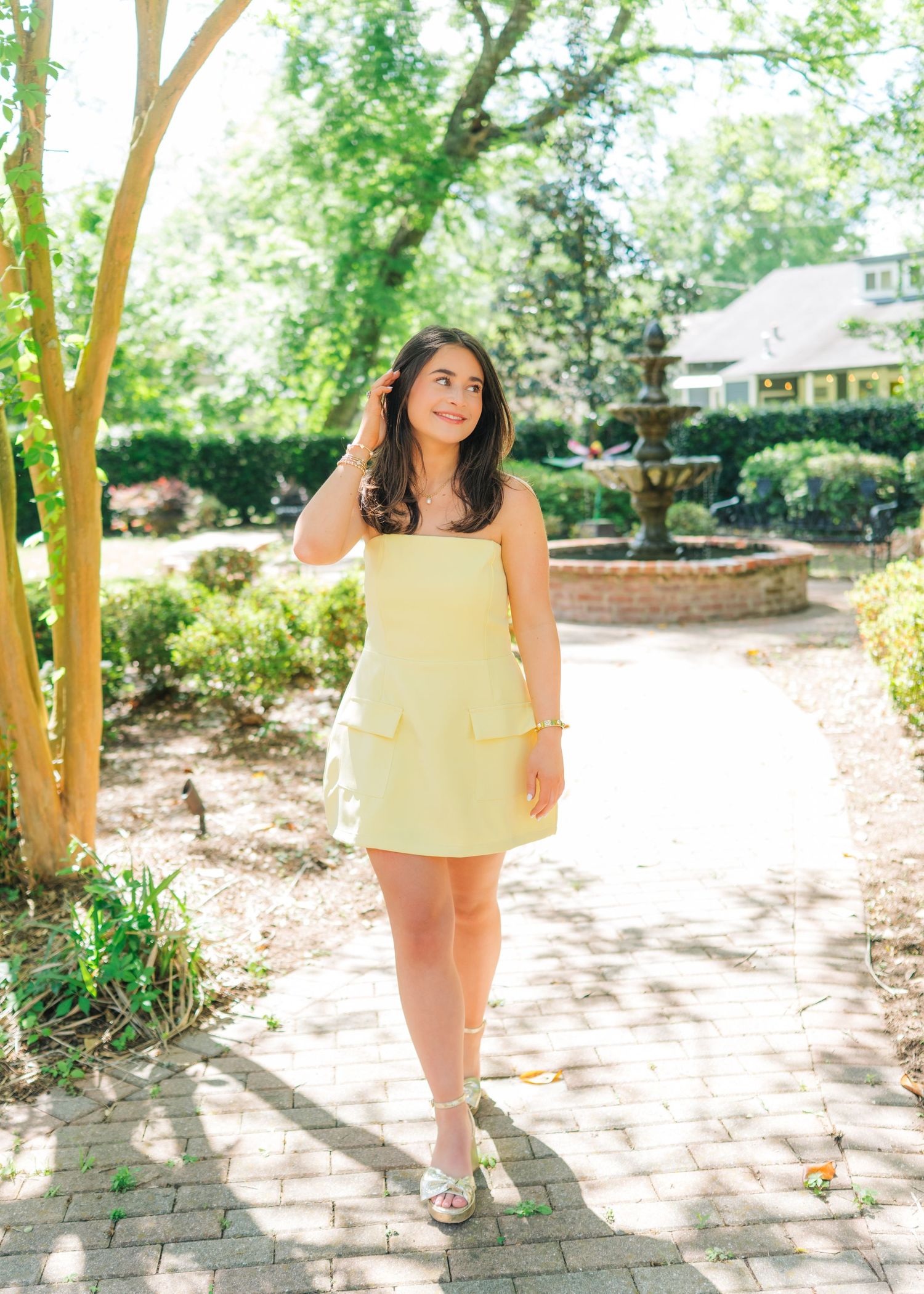Young woman in yellow mini dress poses on brick path in sunny garden with fountain backdrop.