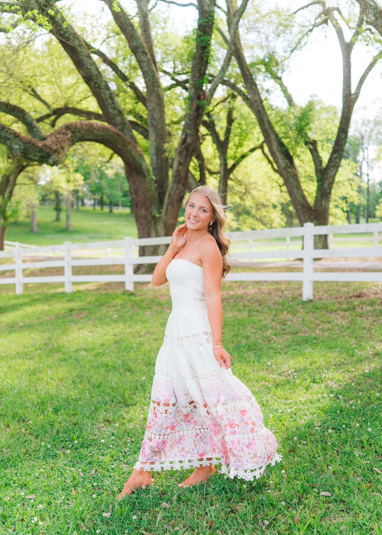 Young woman in floral lace midi dress twirling on green lawn with white fence and oak trees.