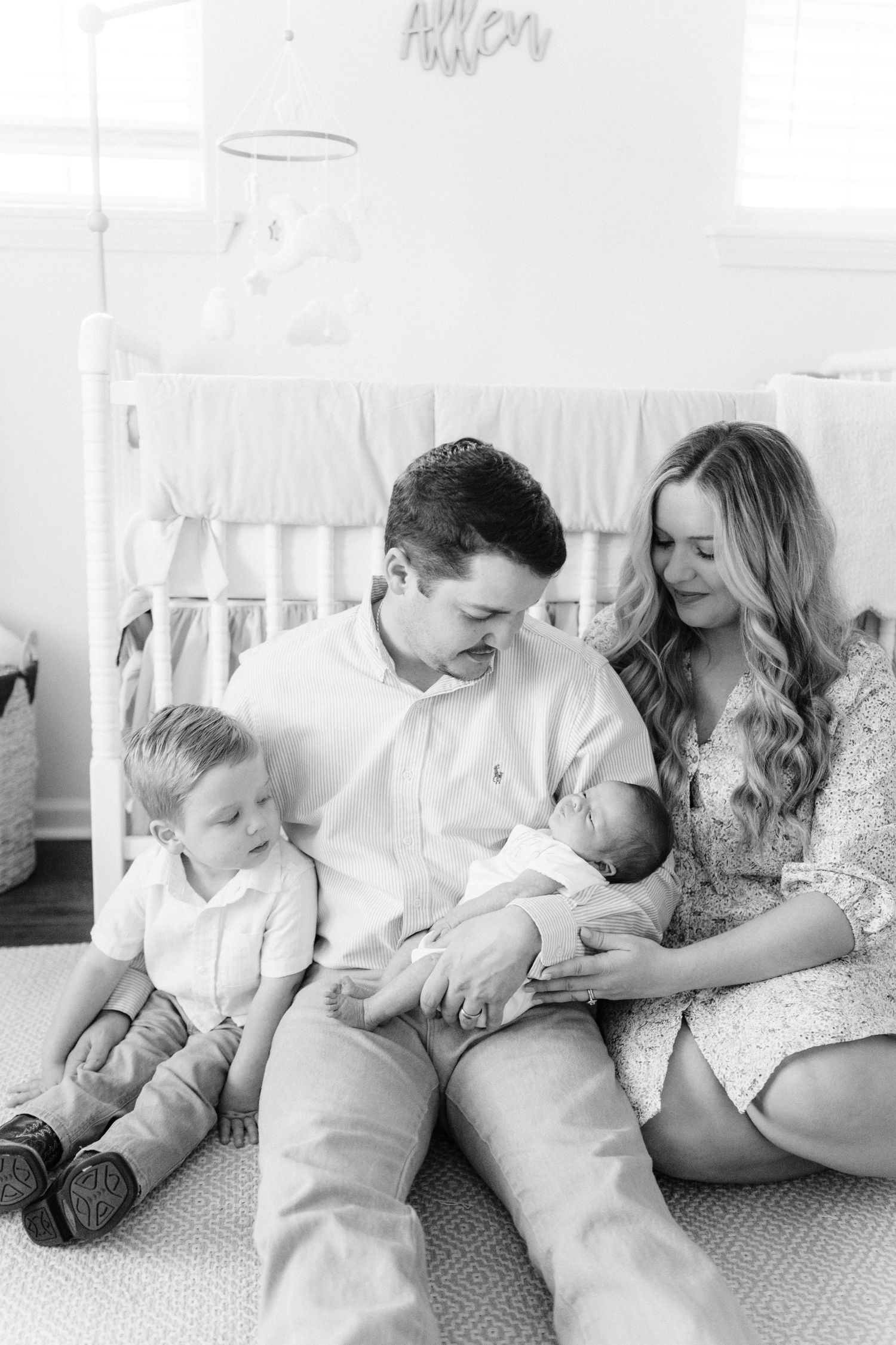 Black and white photo of a family of four sitting on nursery floor, parents admiring their newborn baby.