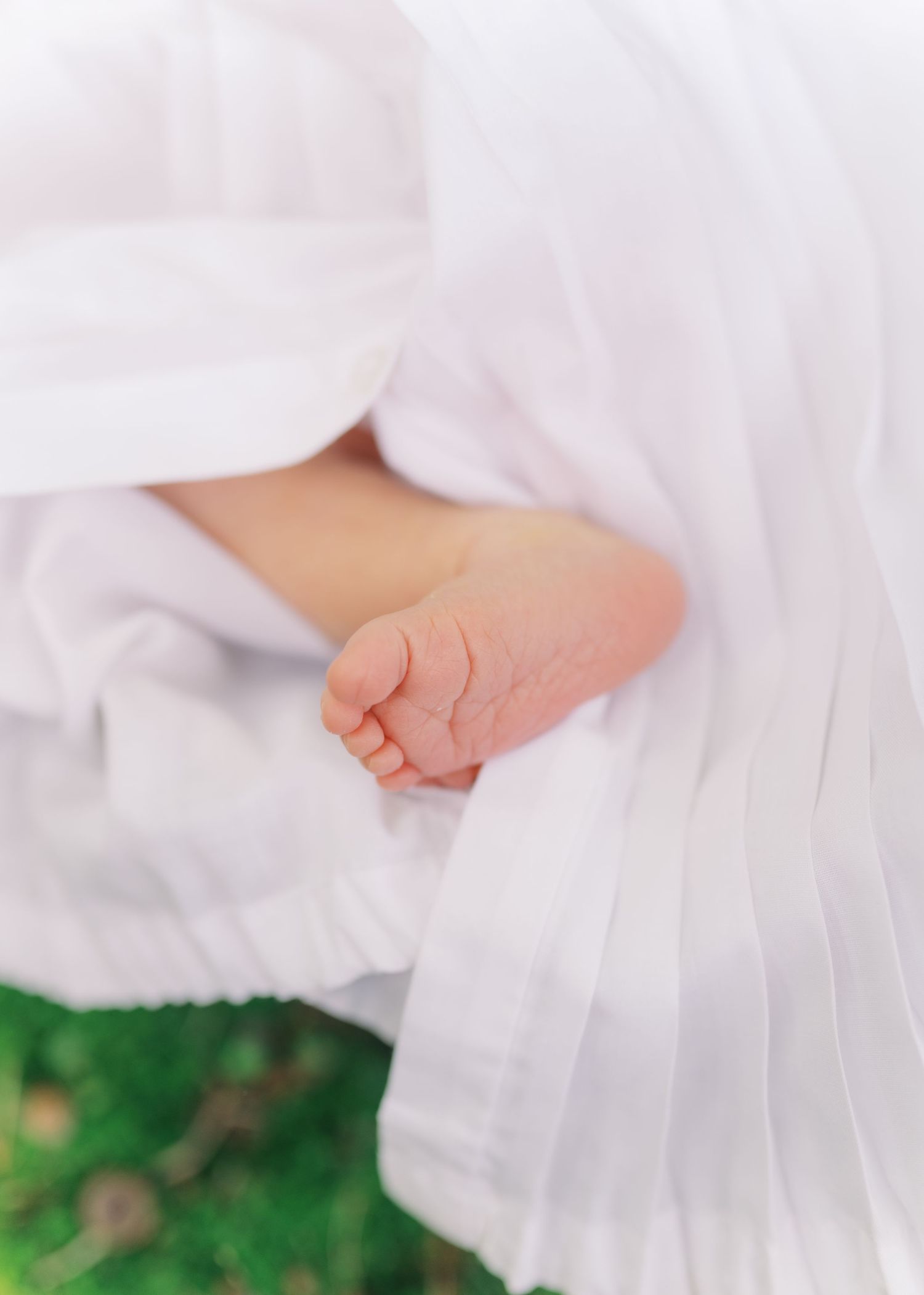 Close-up of tiny newborn baby foot peeking out from soft white swaddle blanket outdoors.