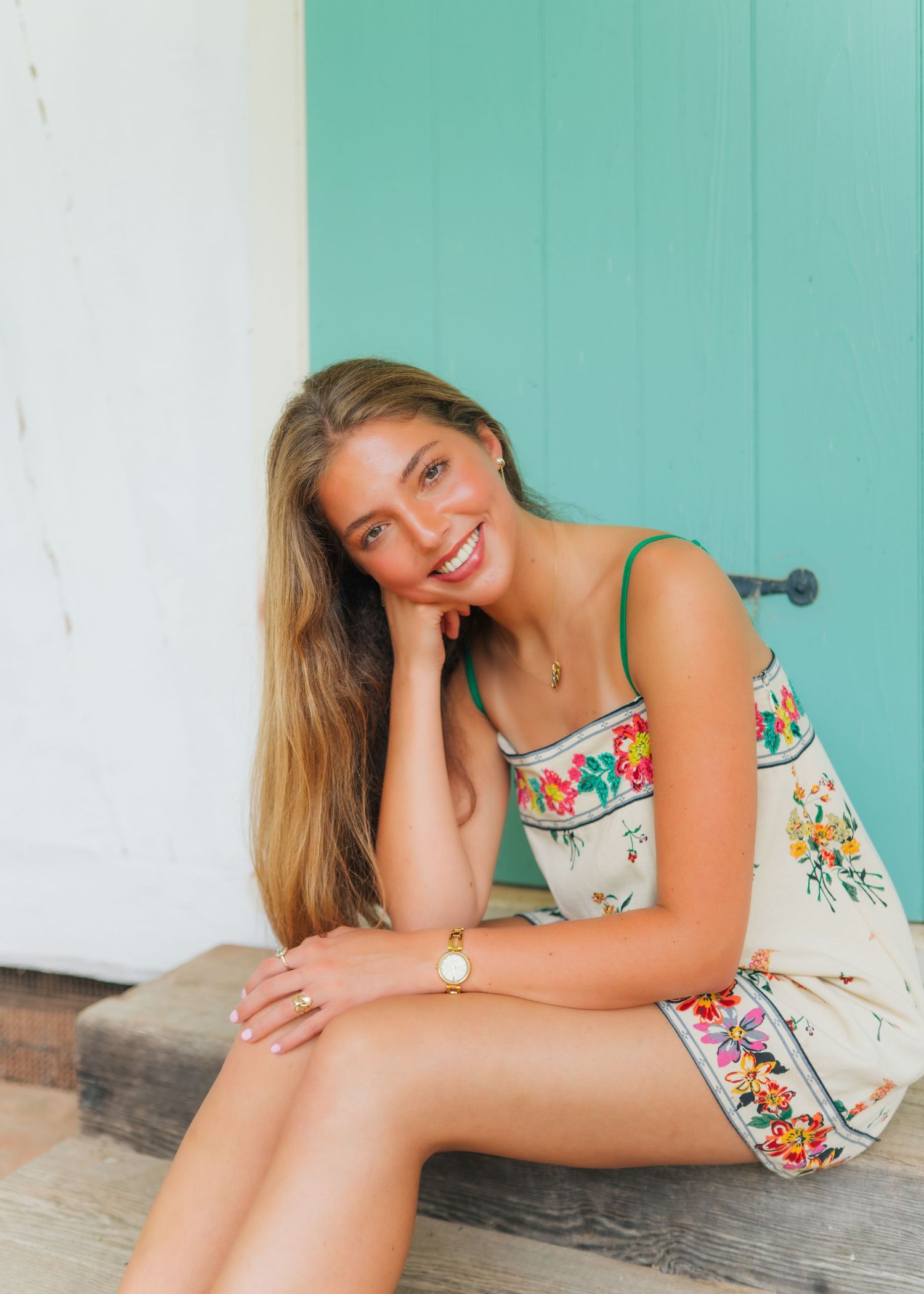 Young woman in floral sundress sitting on steps, smiling, with teal door backdrop.