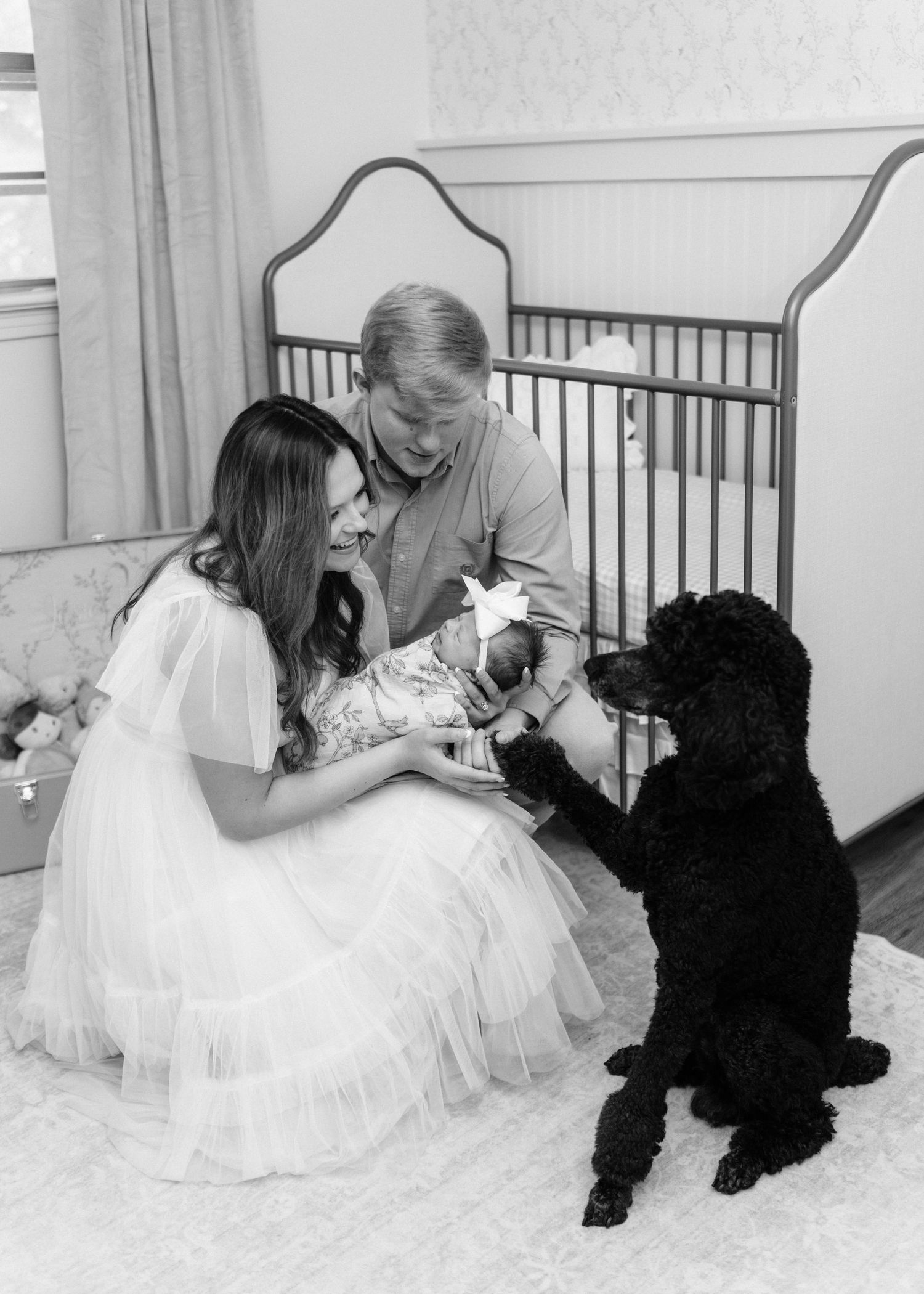 Black and white newborn photo of parents with baby and black poodle by crib.