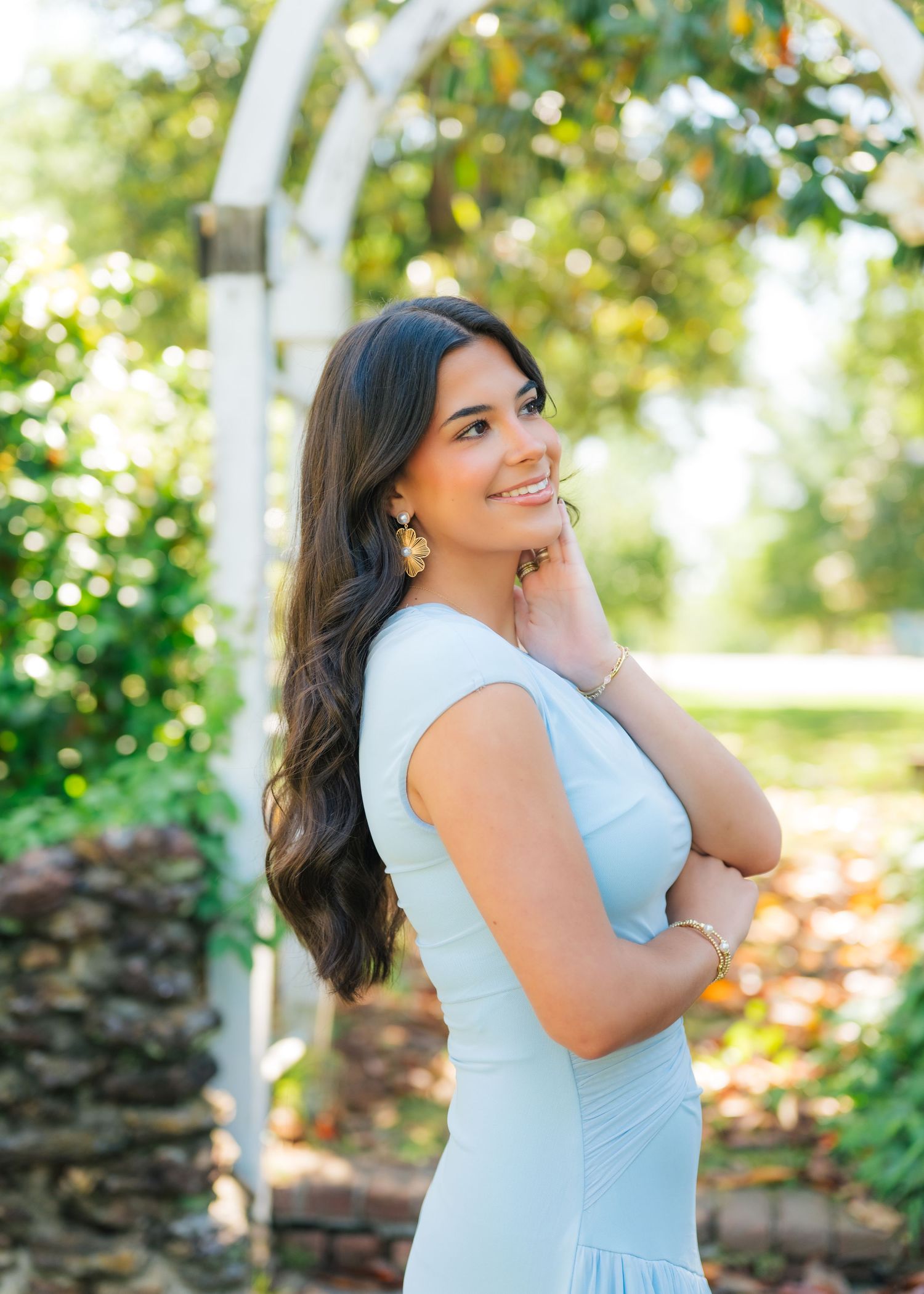 Young woman in light blue dress smiling in a garden with white arbor and lush greenery backdrop.