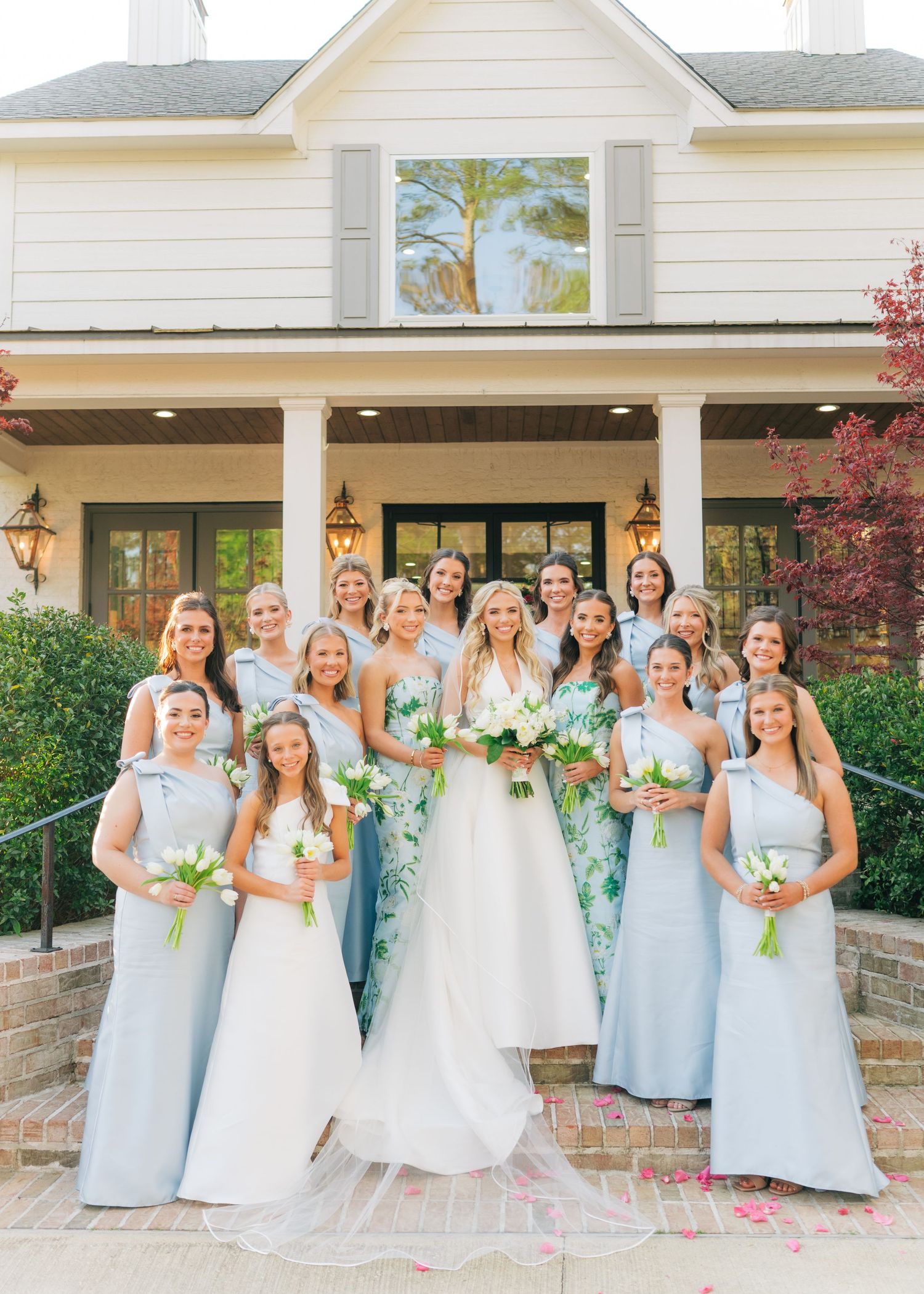 Bride poses with bridesmaids in light blue gowns holding white bouquets in front of elegant white venue.