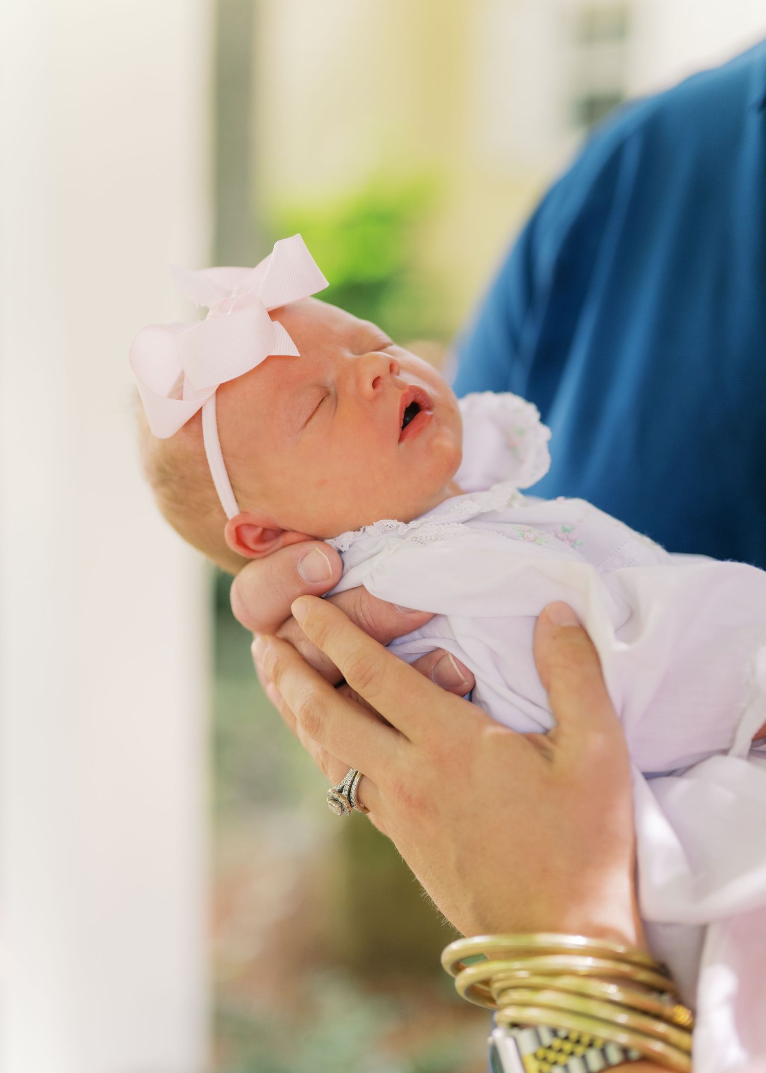 Newborn baby girl in white outfit and pink bow headband being cradled in adult hands outdoors.