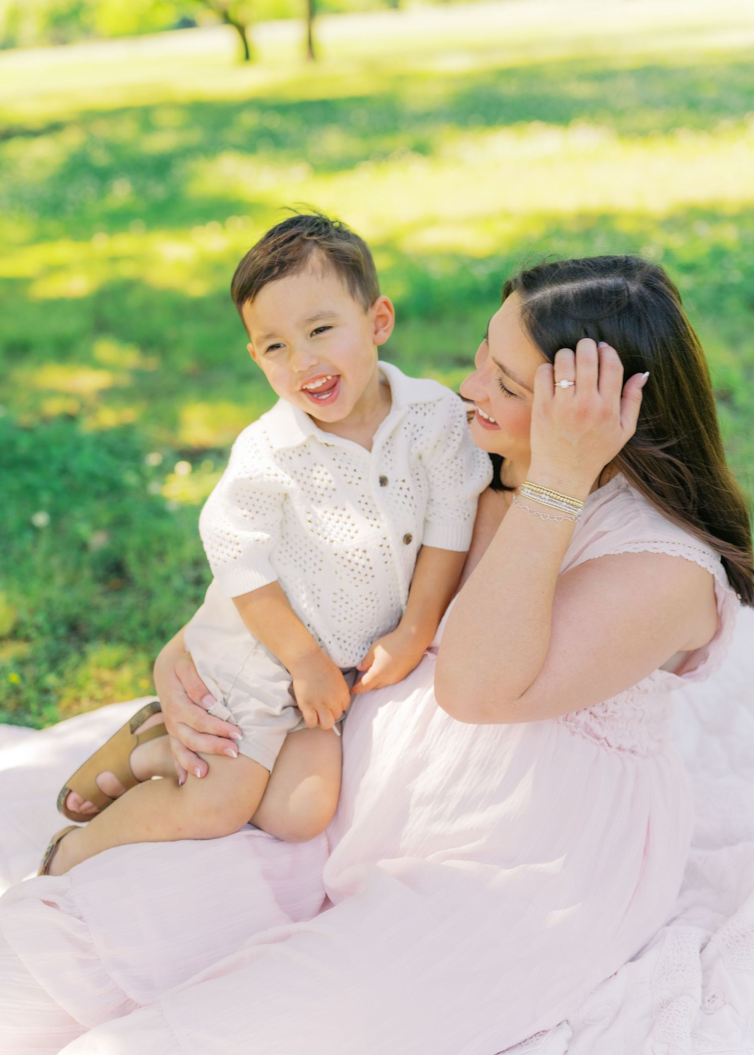 A joyful toddler boy in white sits on his mother's lap outdoors on a blanket in a sunny green park.