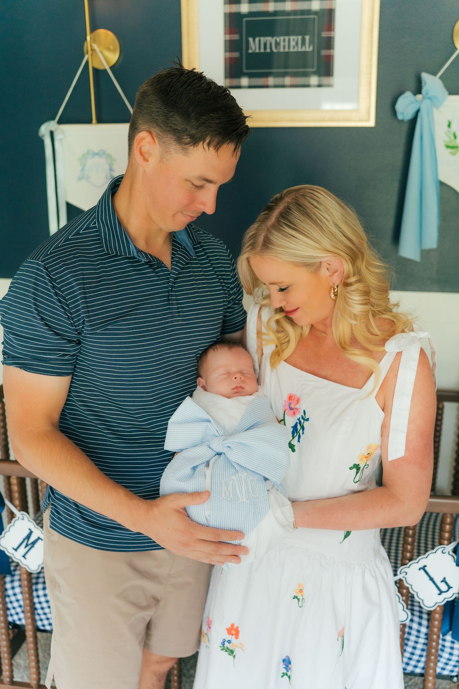 Happy new parents holding their swaddled newborn in a navy nursery decorated with the name Mitchell on the wall.