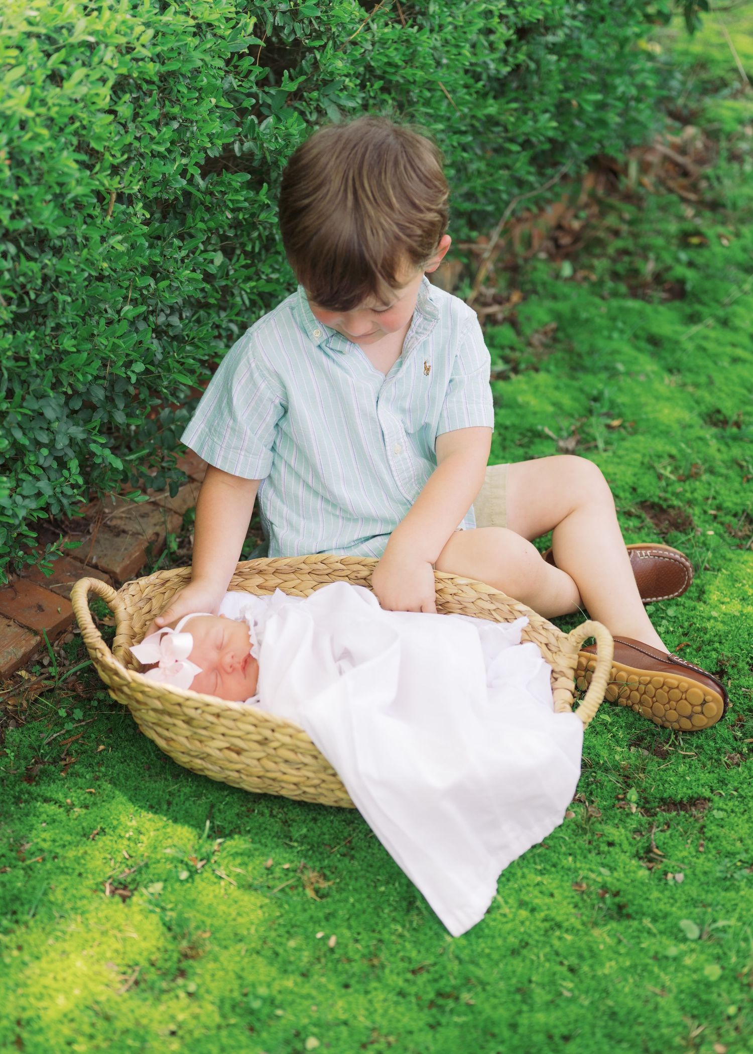 Young boy sitting cross-legged on grass lovingly watching over newborn sister in wicker basket.
