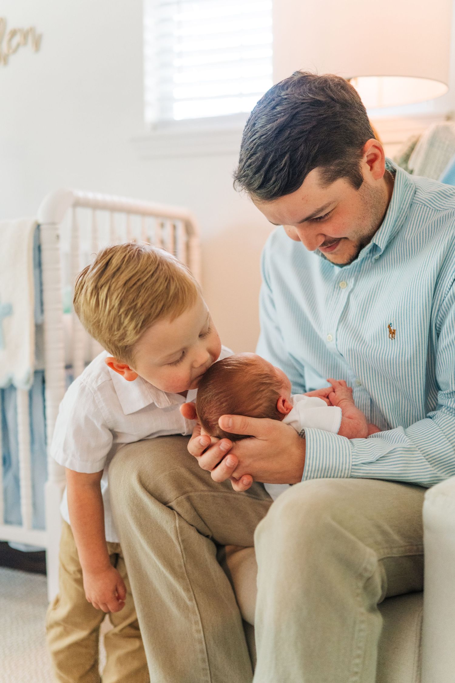 Loving toddler boy kisses newborn sibling held by father in a cozy nursery setting.