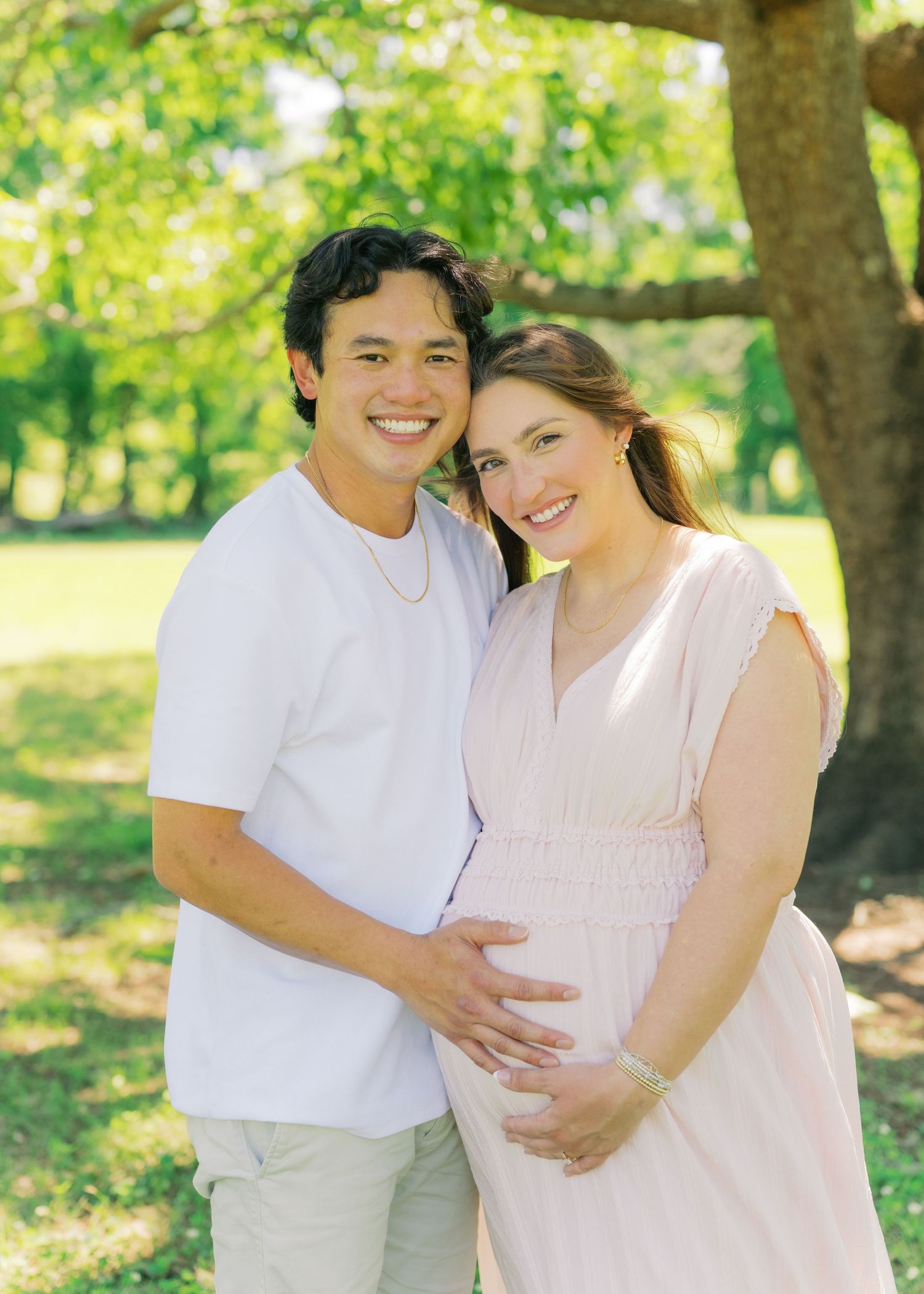 Smiling couple outdoors, pregnant woman in pink dress cradling baby bump with partner in white shirt.