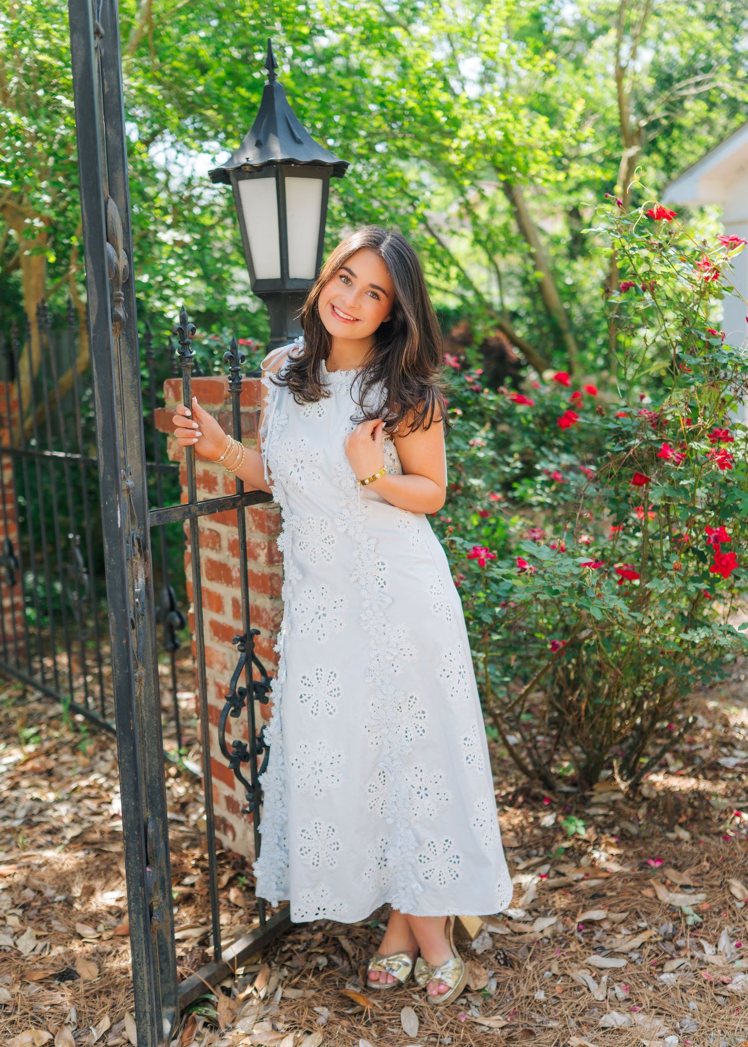 Young woman in white eyelet maxi dress standing by an iron gate with red roses in the background.