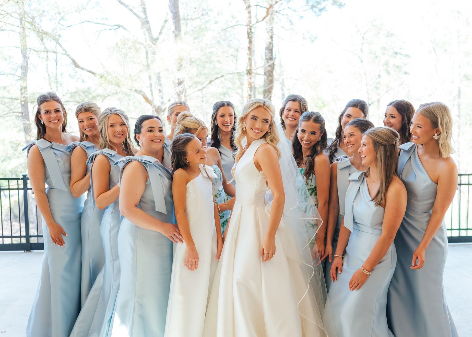 Bride in white gown surrounded by smiling bridesmaids in light blue dresses at outdoor wedding.