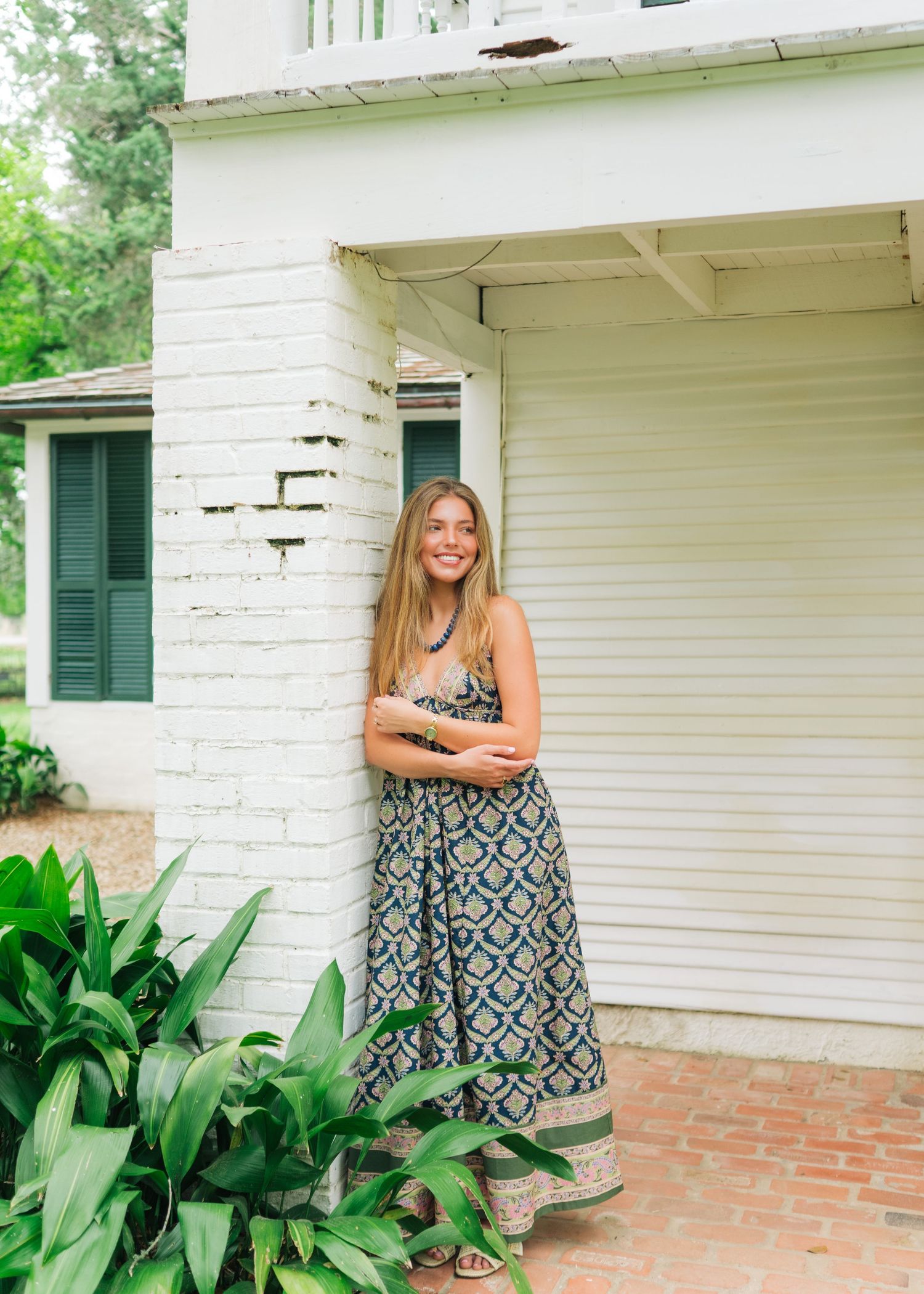 Smiling young woman in floral maxi dress posing beside white brick column of historic Southern home.