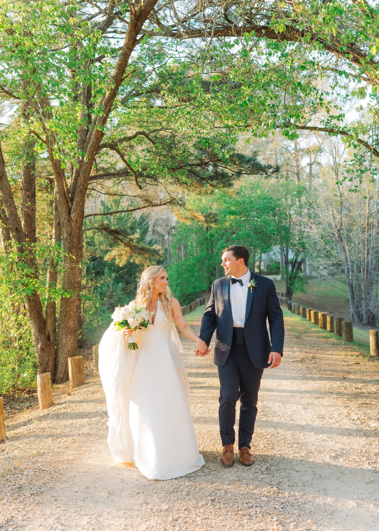 Bride and groom walk hand in hand down a sunlit tree-lined path, bride holding a white bouquet.