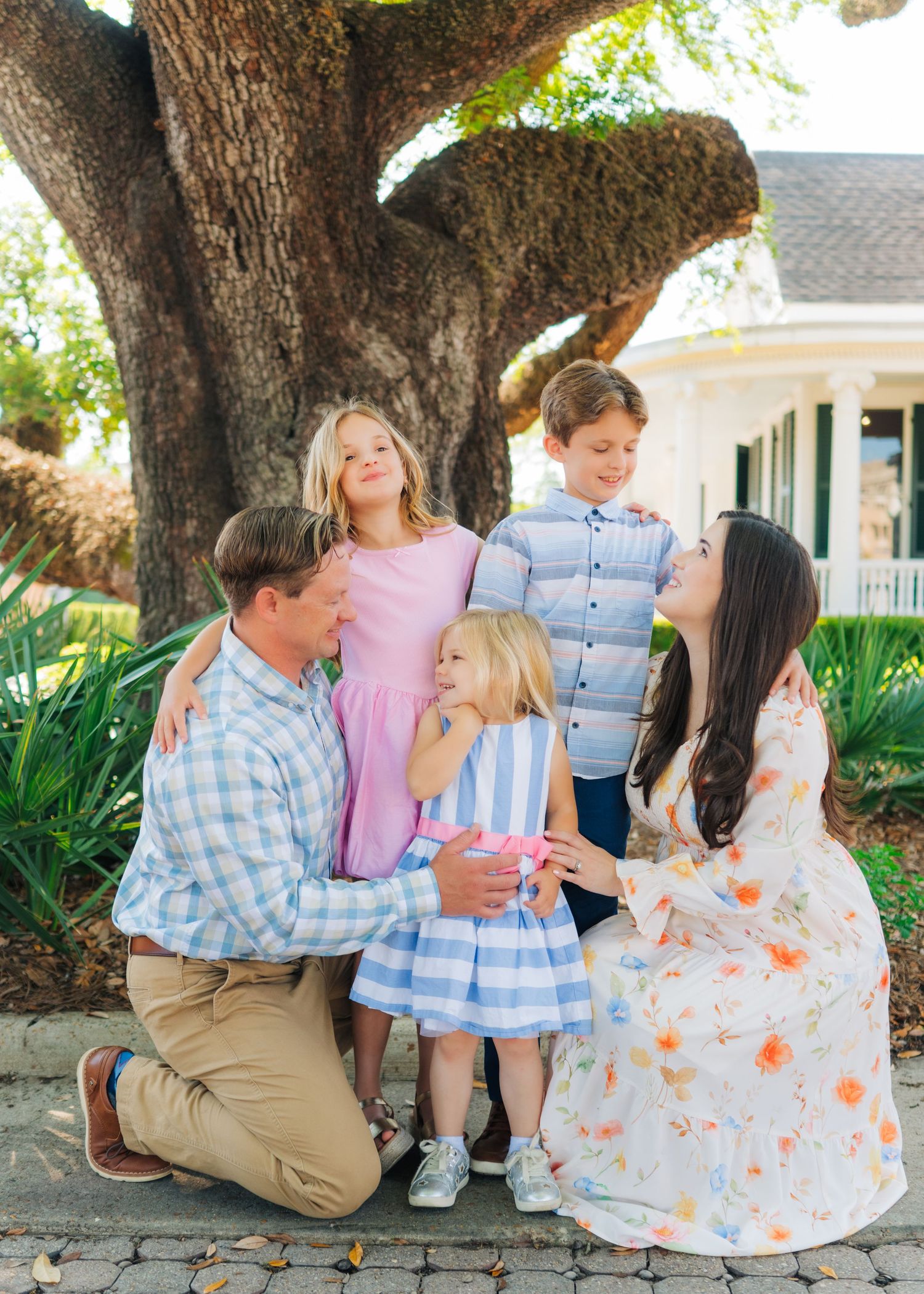 A family of five poses under a large oak tree, with parents and three young children dressed in pastel spring outfits.