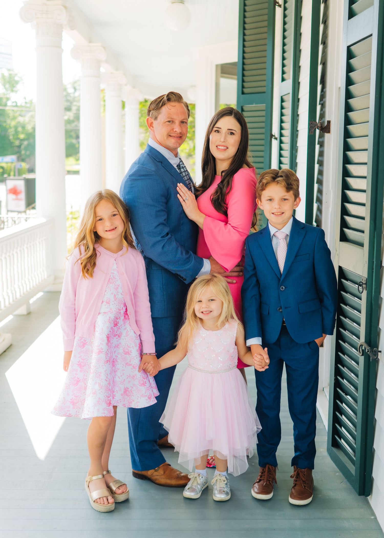 A family of five poses on a porch, parents with three children dressed in coordinating blue and pink formal outfits.