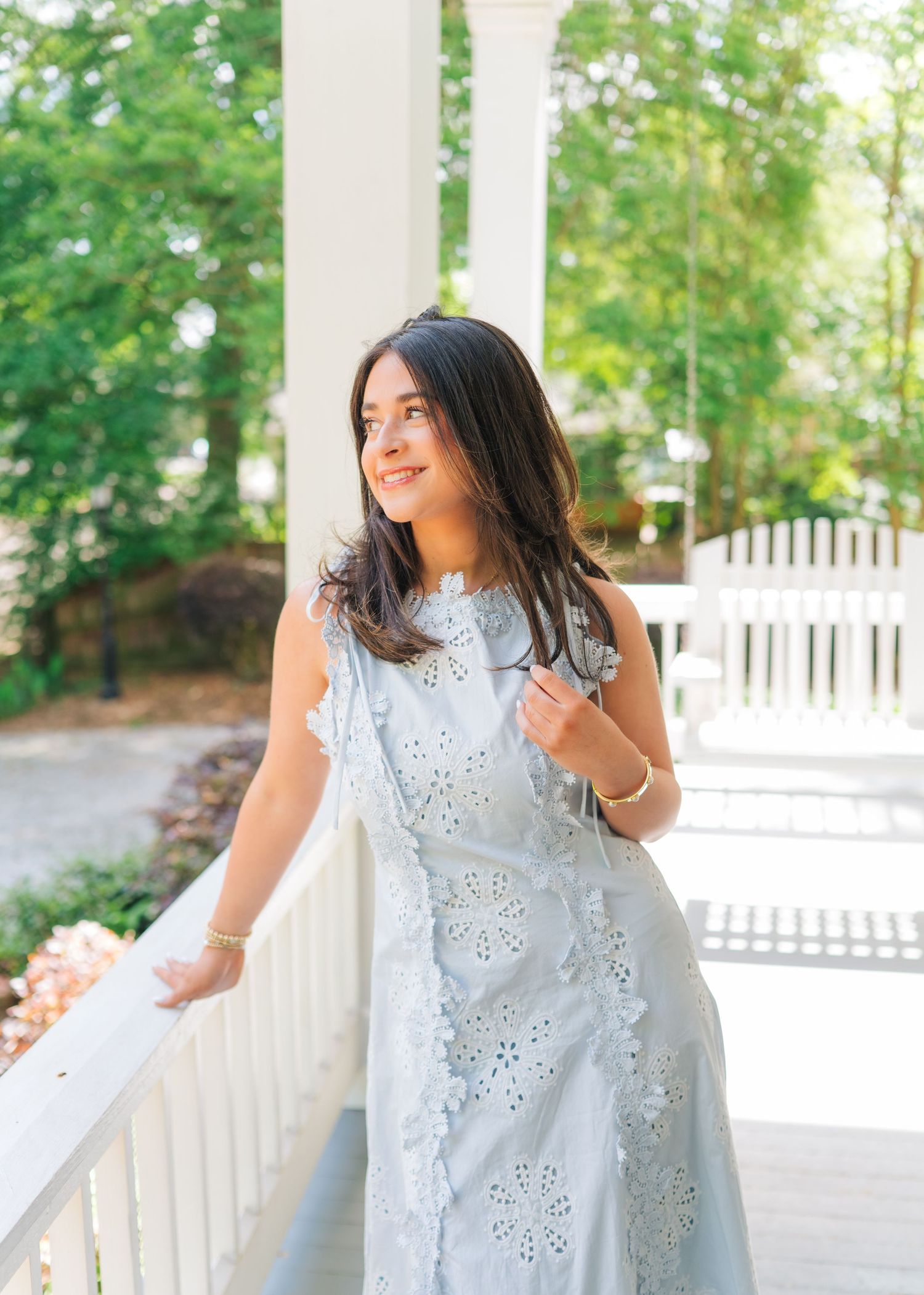 Young woman in light blue eyelet dress smiling on a white porch with lush green trees in background.