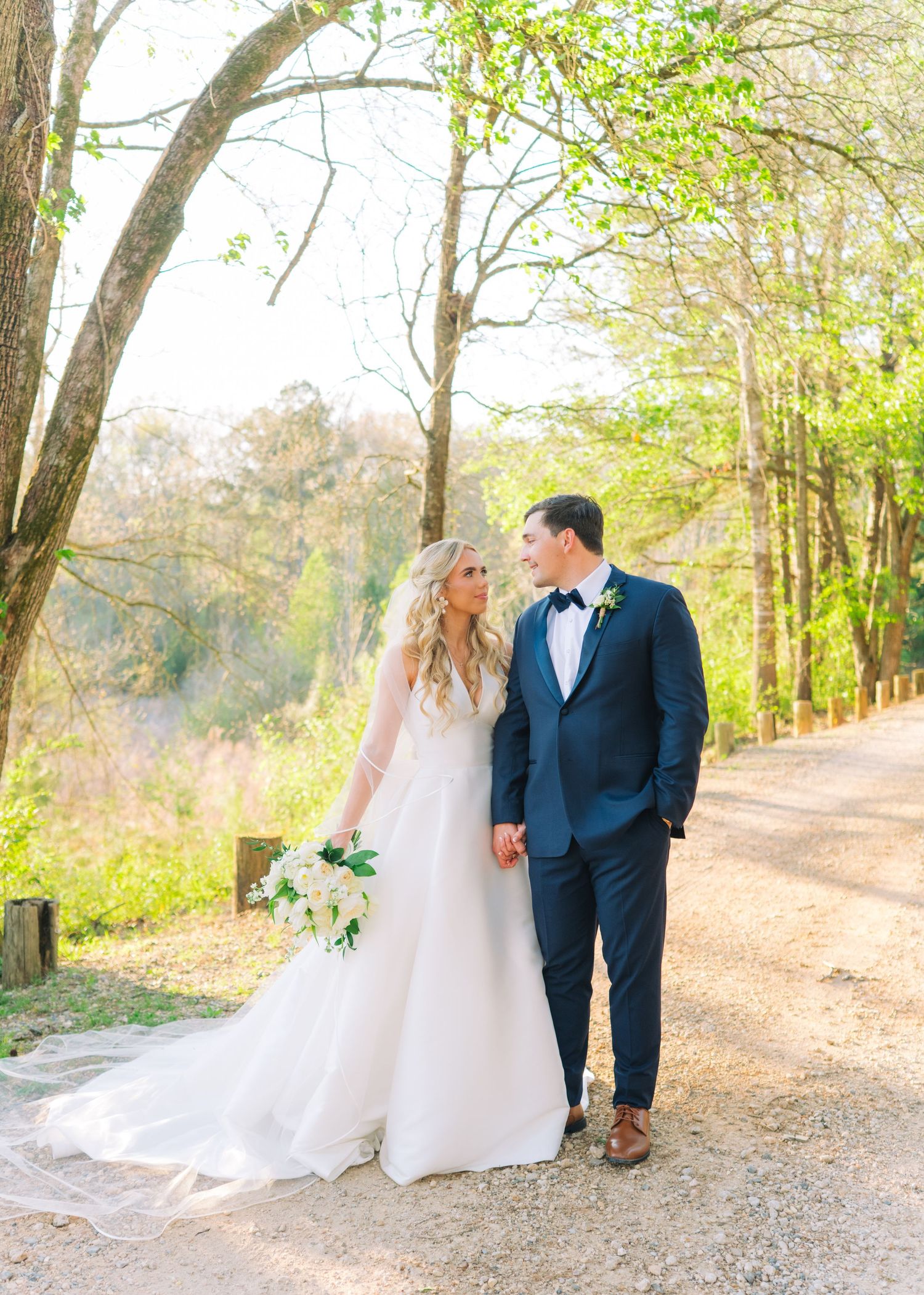 Bride and groom walk hand in hand along a tree-lined path in spring wedding attire.