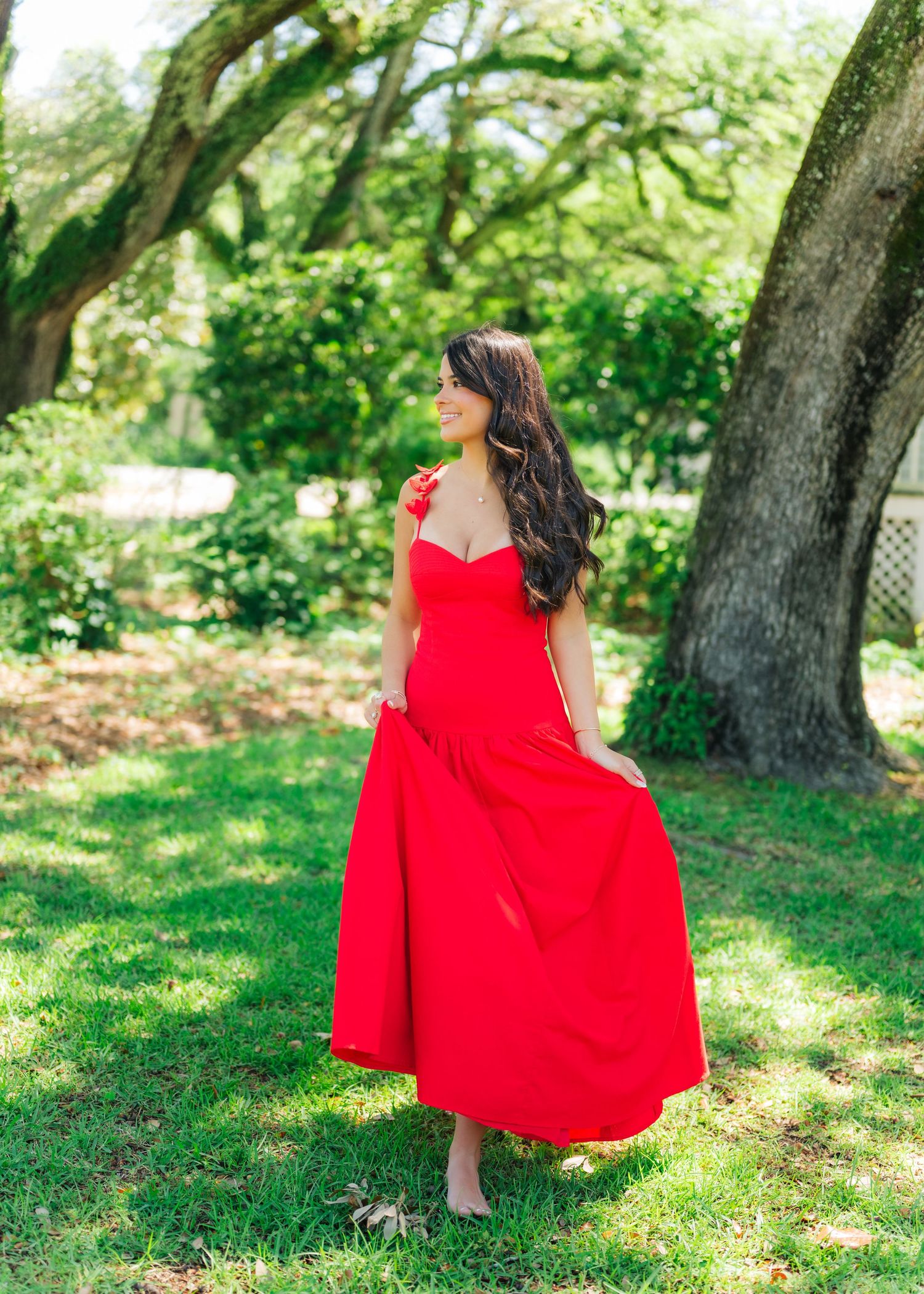 Woman in flowing red maxi dress standing gracefully outdoors under large oak trees on lush green grass.