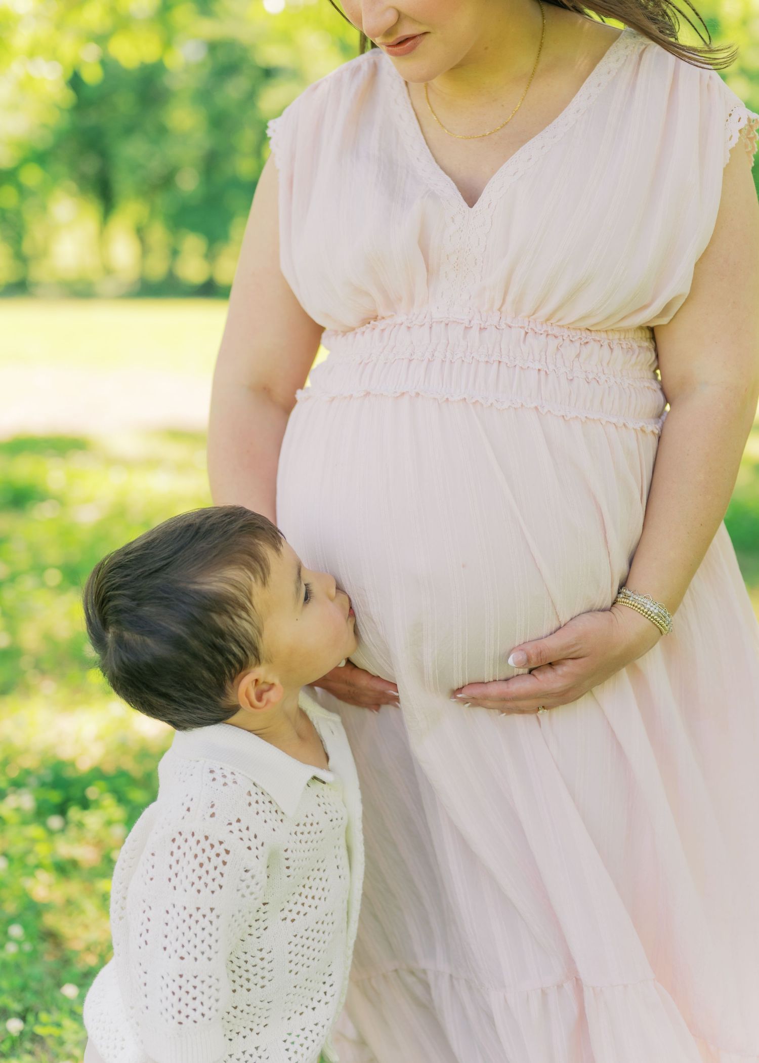 A young boy kisses a pregnant woman's belly outdoors, both dressed in soft whites and pink.