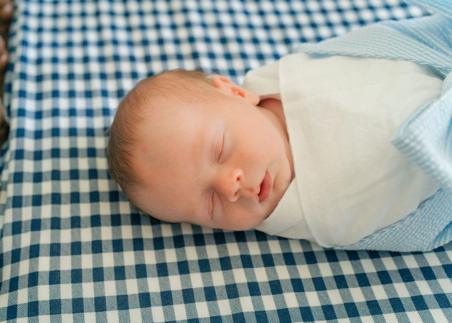 Peaceful newborn baby swaddled in white blanket sleeping on blue gingham fabric.