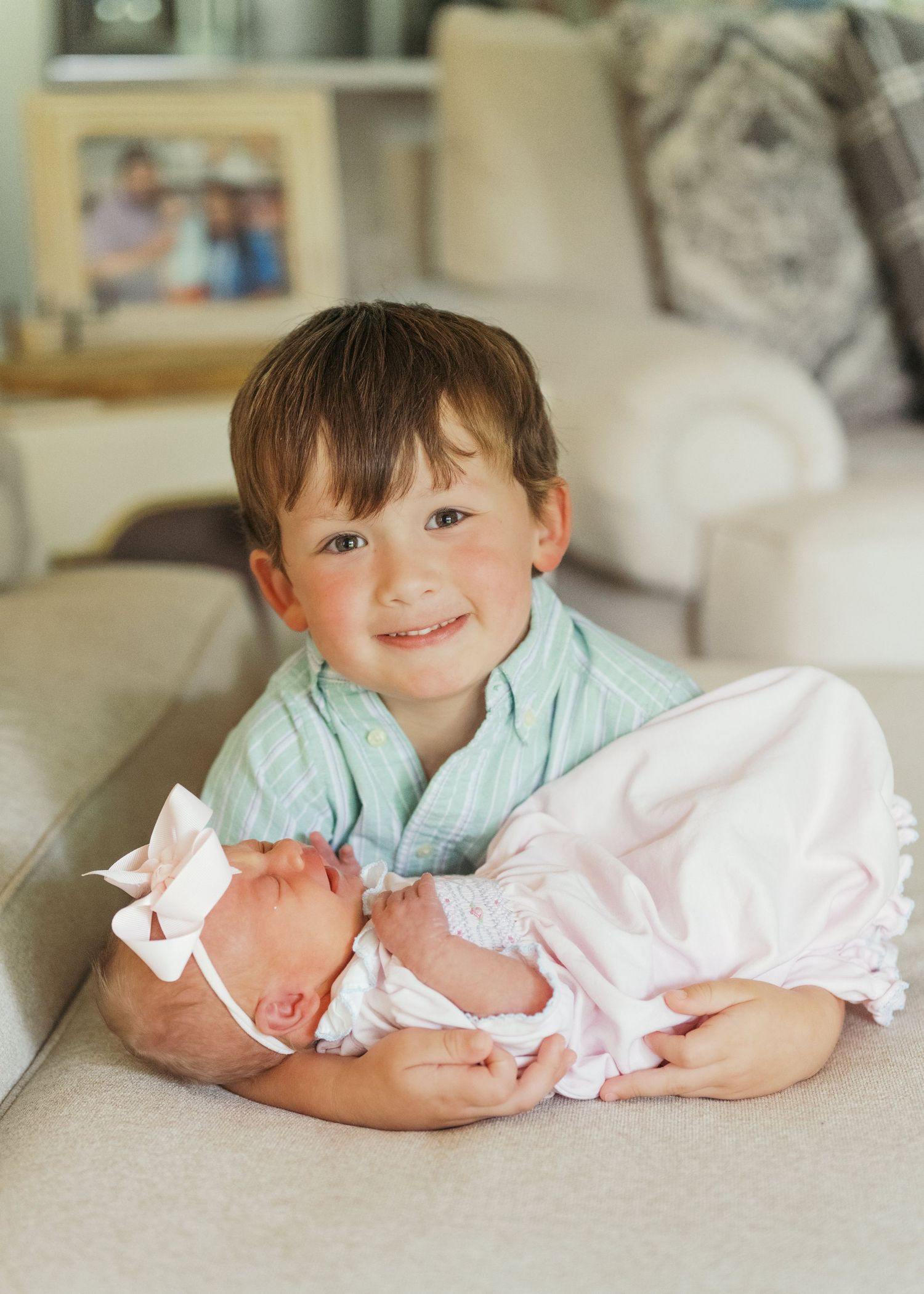 Smiling young boy holds sleeping newborn baby girl in pink outfit with bow headband on a couch indoors.
