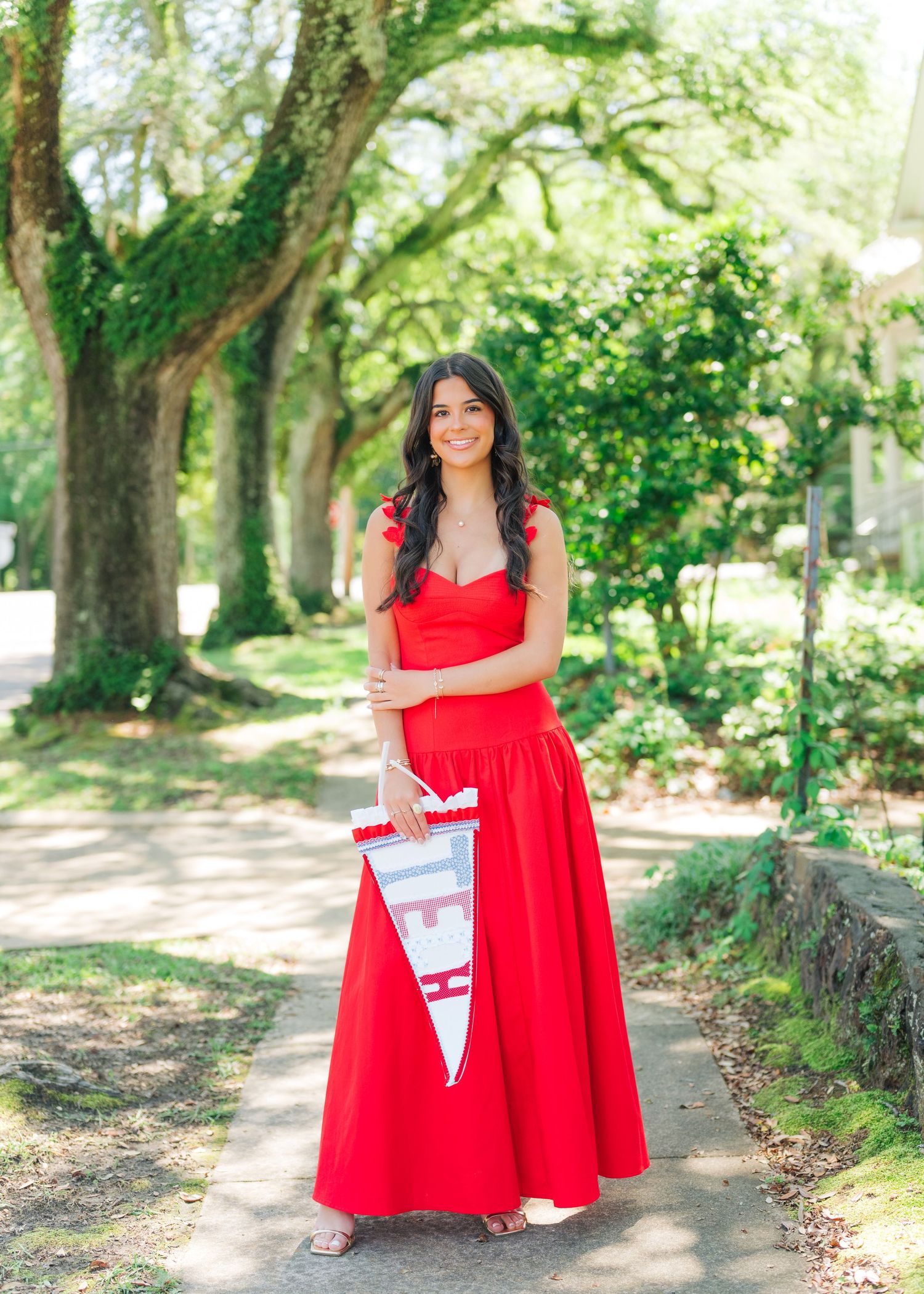 Young woman in red maxi dress holding a pennant flag, posing on a tree-lined path outdoors.