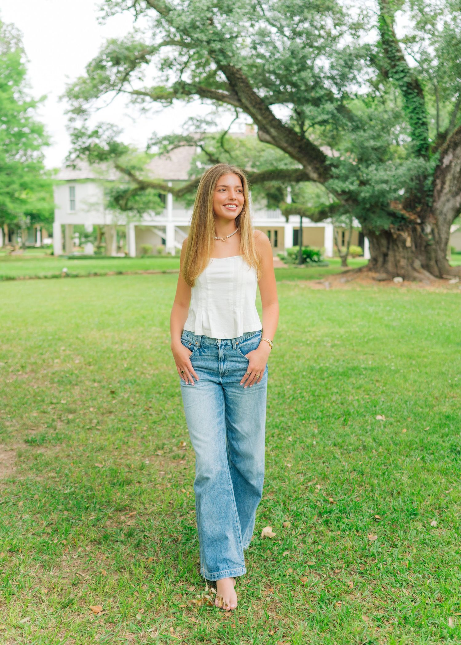 Smiling young woman in white top and wide-leg jeans standing on green lawn with large oak tree.