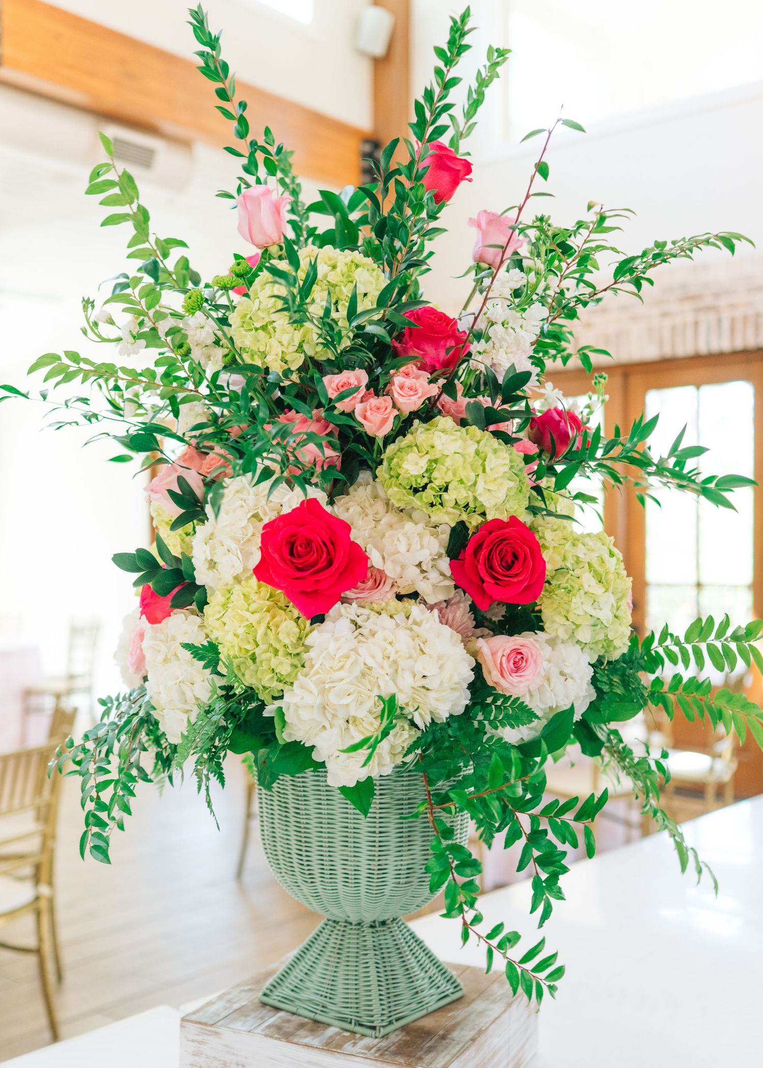 Lush wedding centerpiece featuring red roses, white hydrangeas, and greenery in a mint green wicker urn vase.