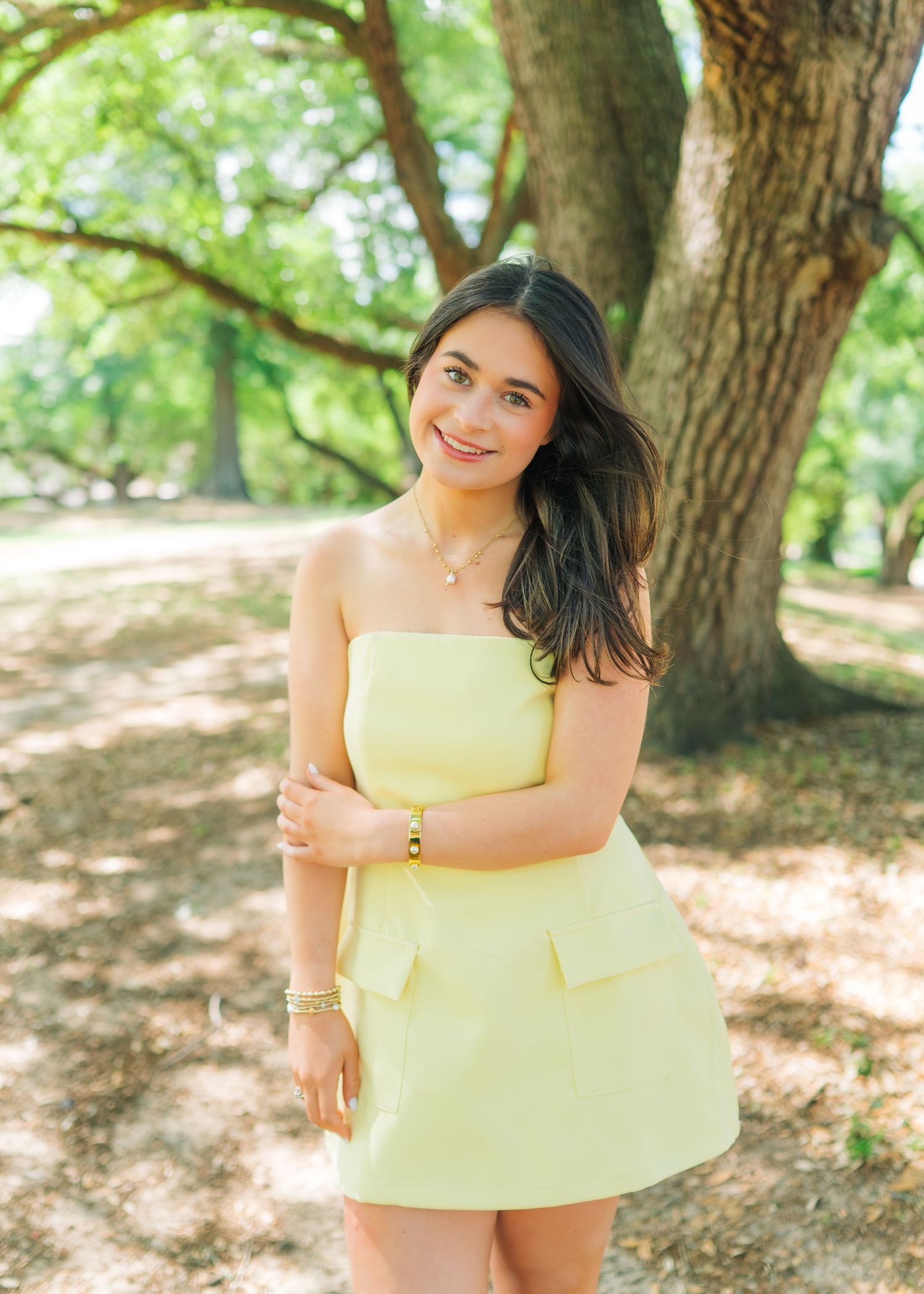 Young woman in a strapless yellow mini dress smiling under a large oak tree in a park.