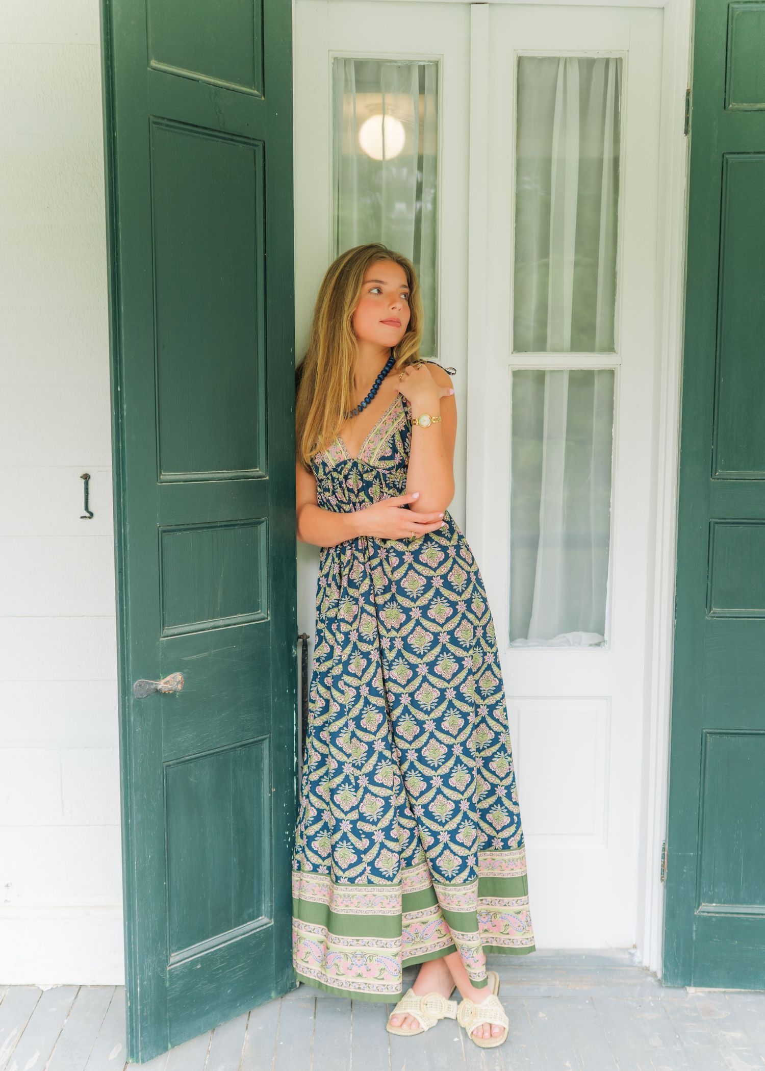 Woman in blue patterned maxi dress standing in green doorway of white building.