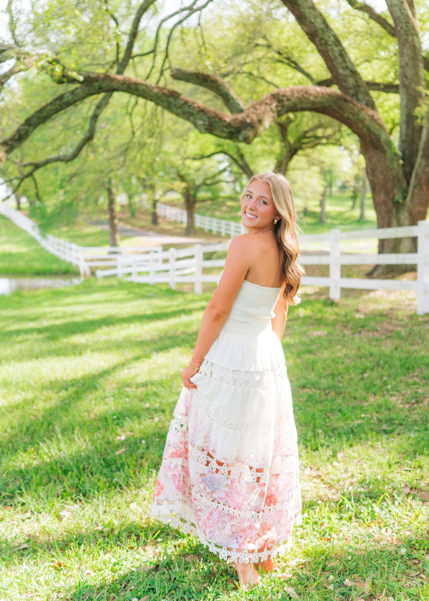 Blonde woman in strapless floral lace dress smiling over shoulder on sunny lawn with oak trees.