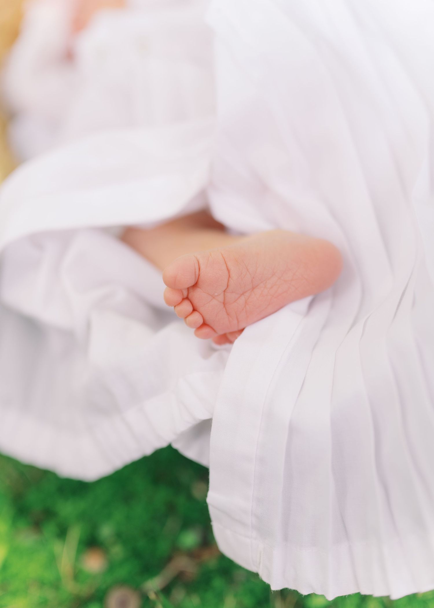 Close-up of a newborn baby's tiny feet peeking out from a delicate white pleated dress outdoors.