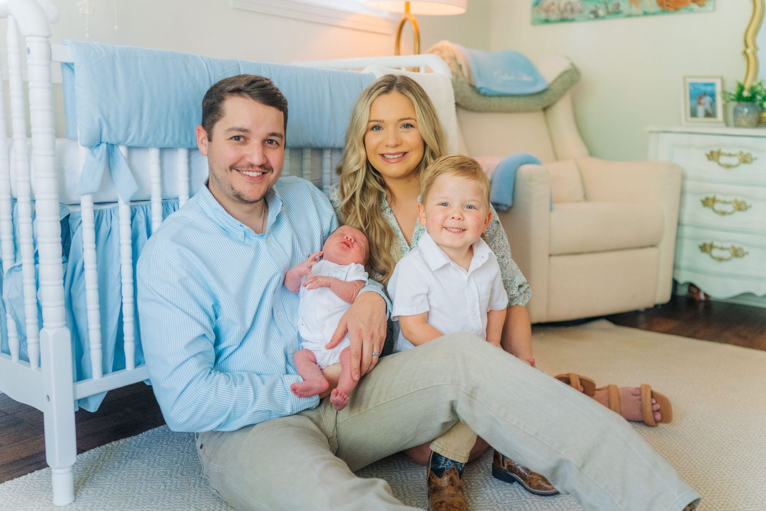 Happy family of four posing in a blue nursery, parents holding newborn baby and toddler son.