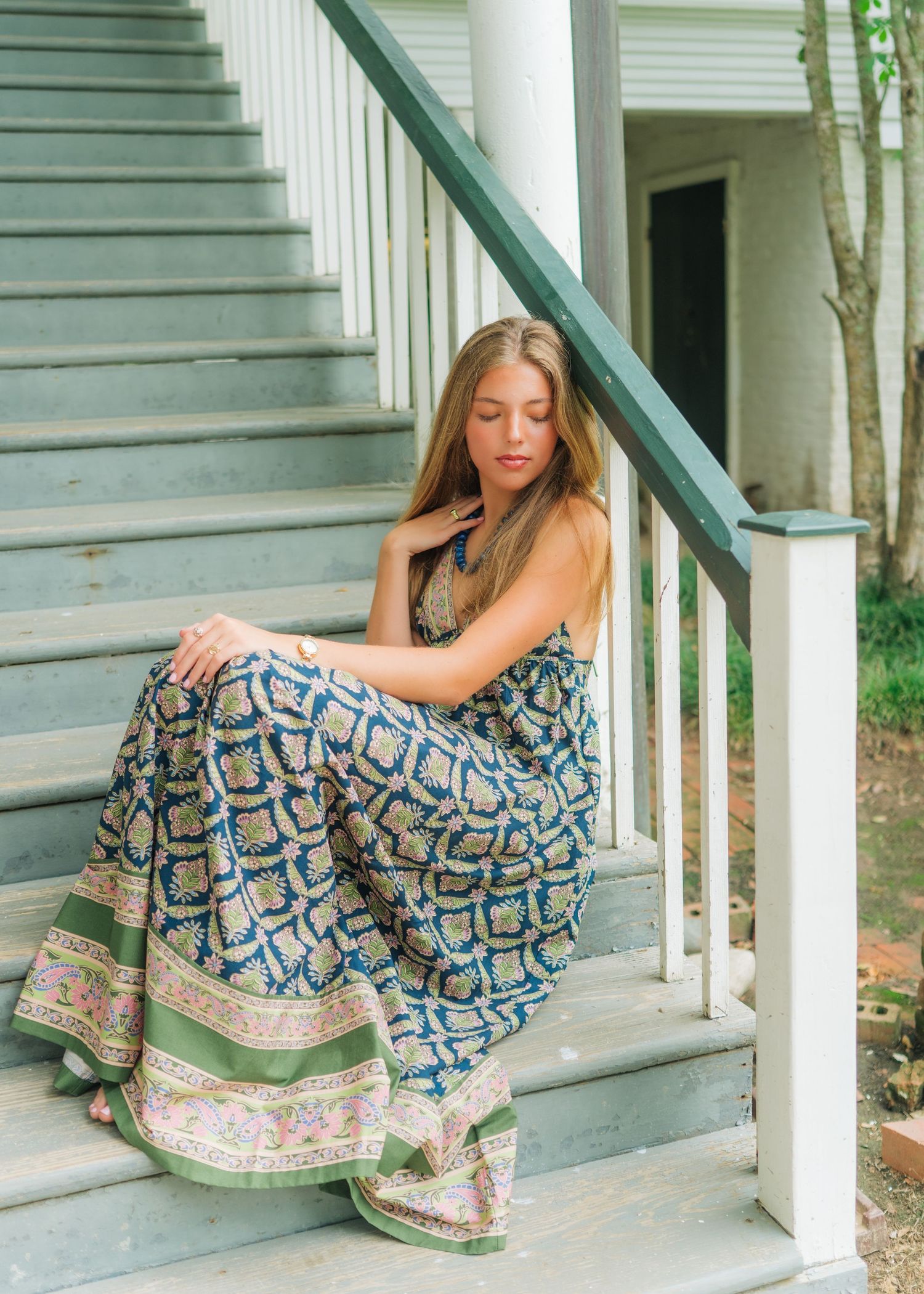 Young woman in floral maxi dress sitting on wooden porch steps outdoors.