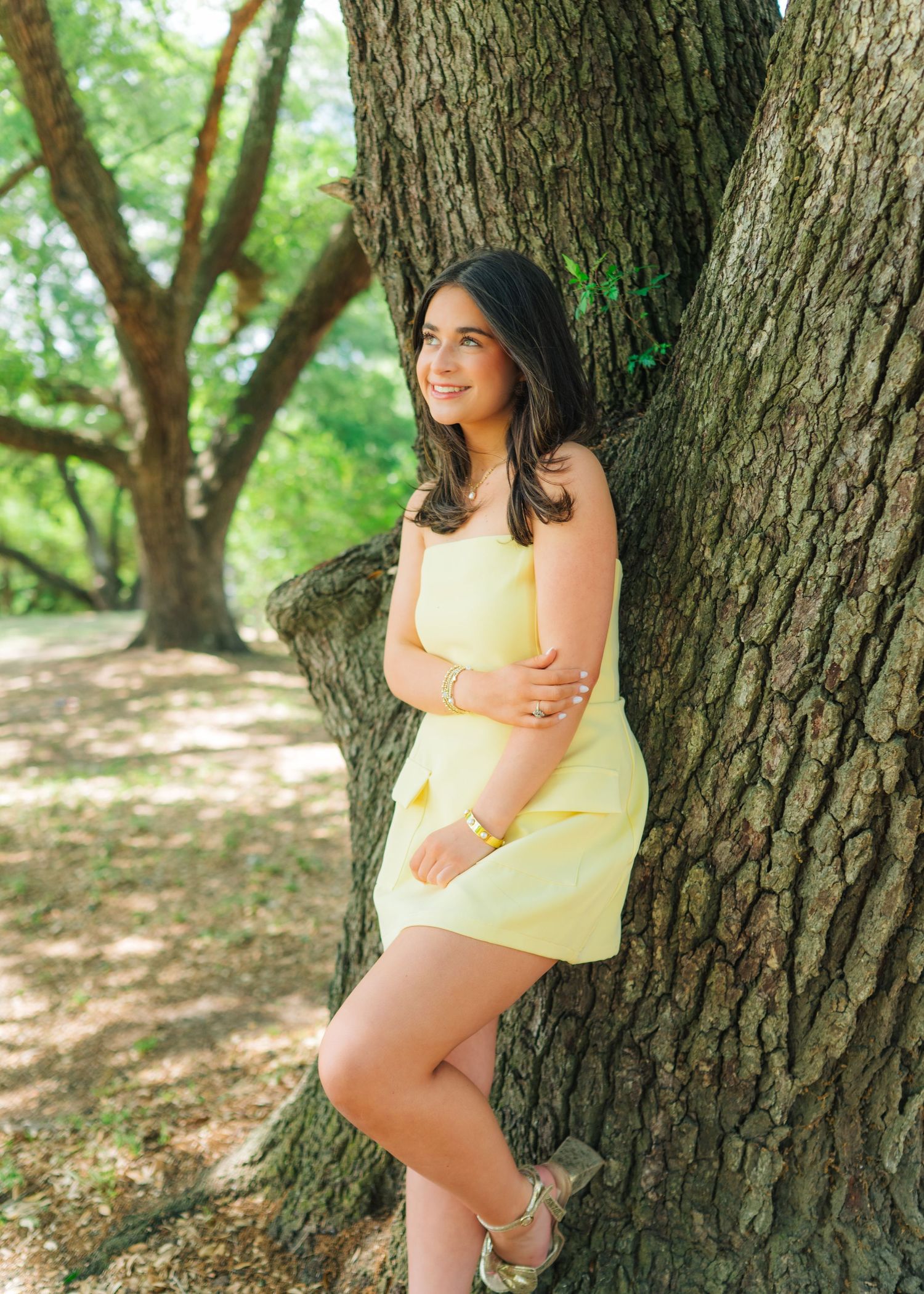 Smiling young woman in yellow mini dress leaning against a large tree trunk in a sunny park.