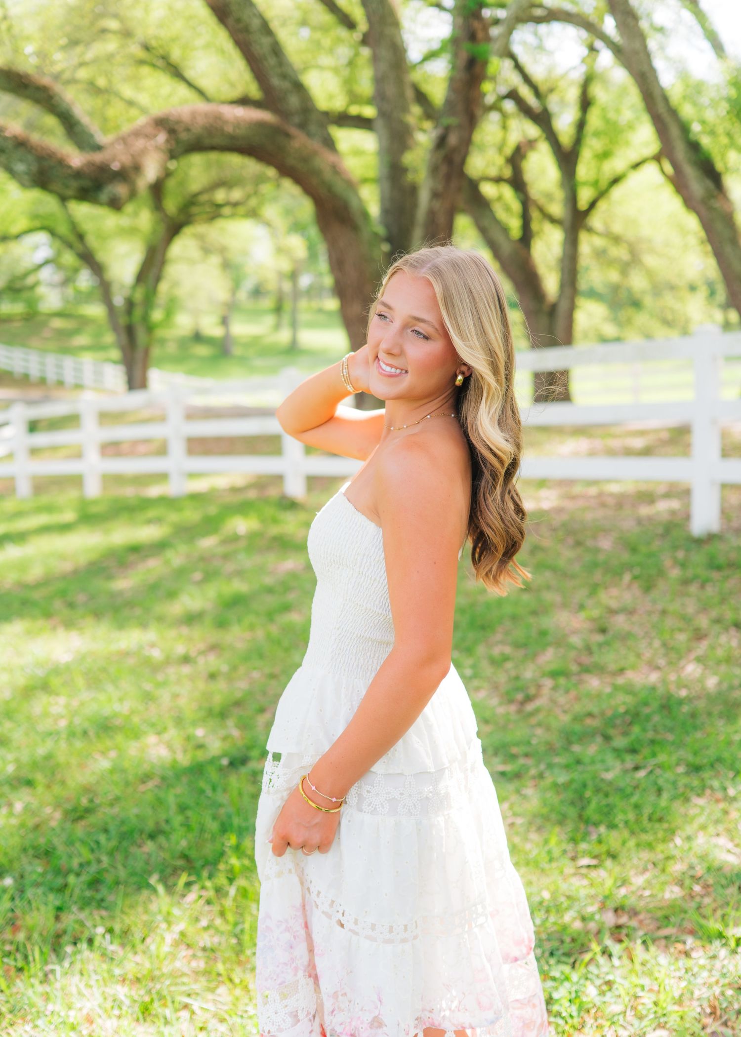 Young woman in white sundress smiling outdoors near white fence with green trees in bright sunlight.