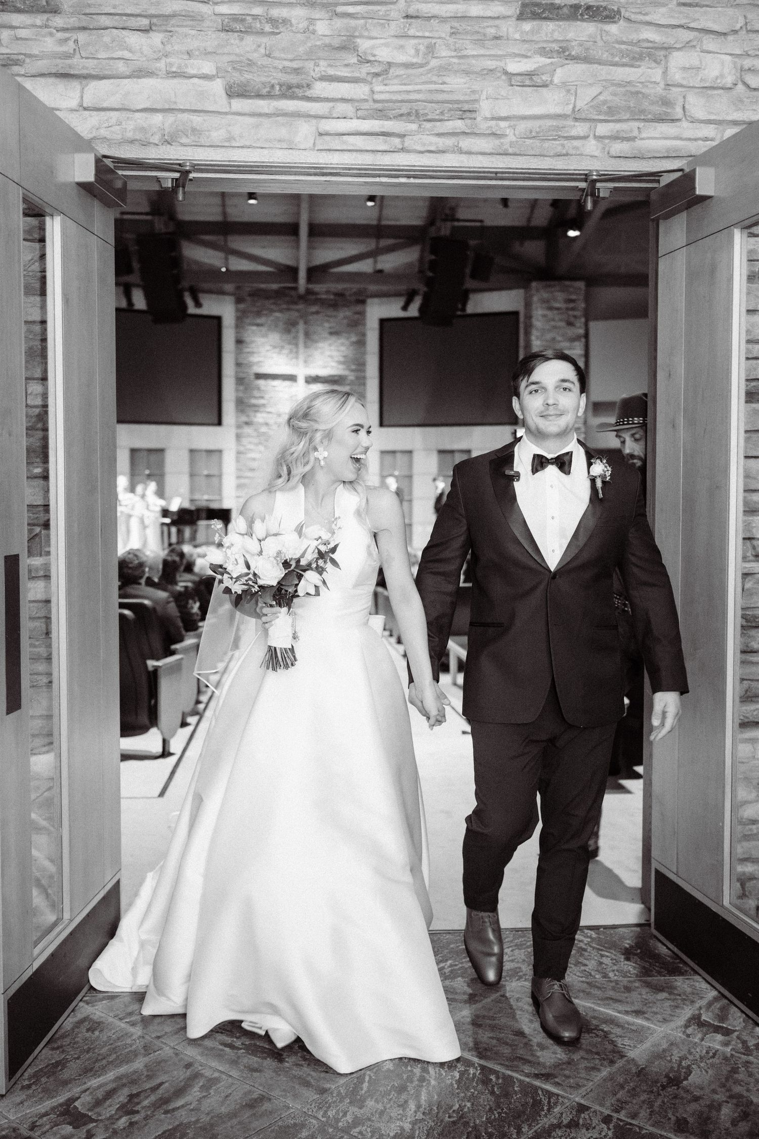 Newlyweds holding hands walk through church doors in black and white wedding portrait.