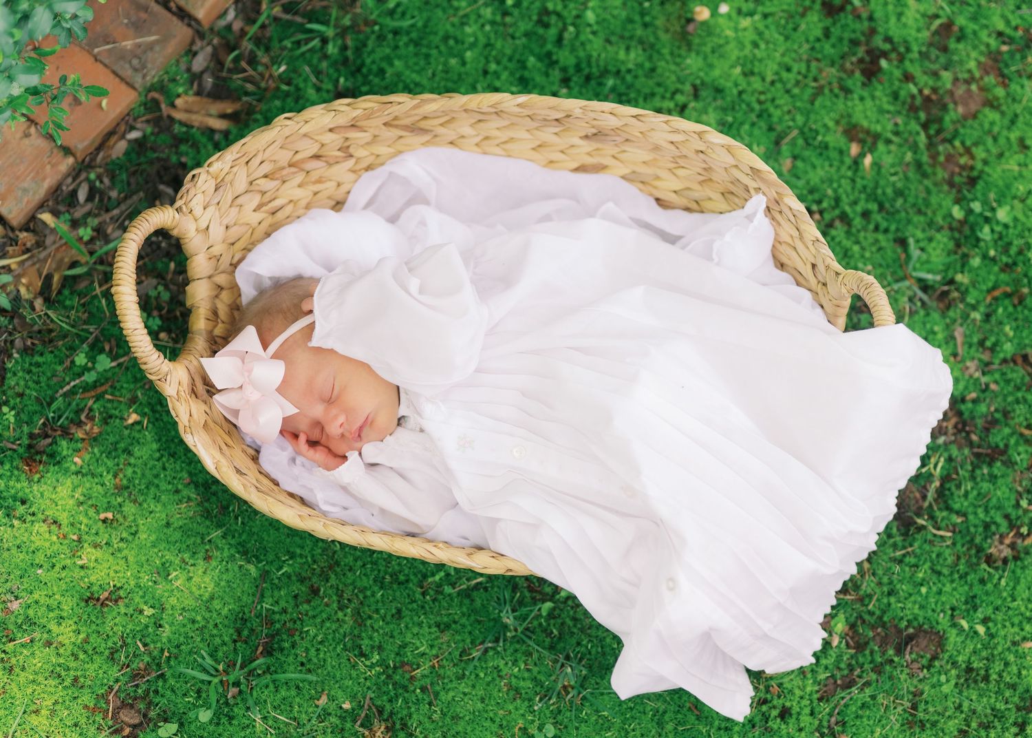 Newborn baby sleeping peacefully in a wicker basket on green grass, wrapped in white fabric with a pink bow headband.