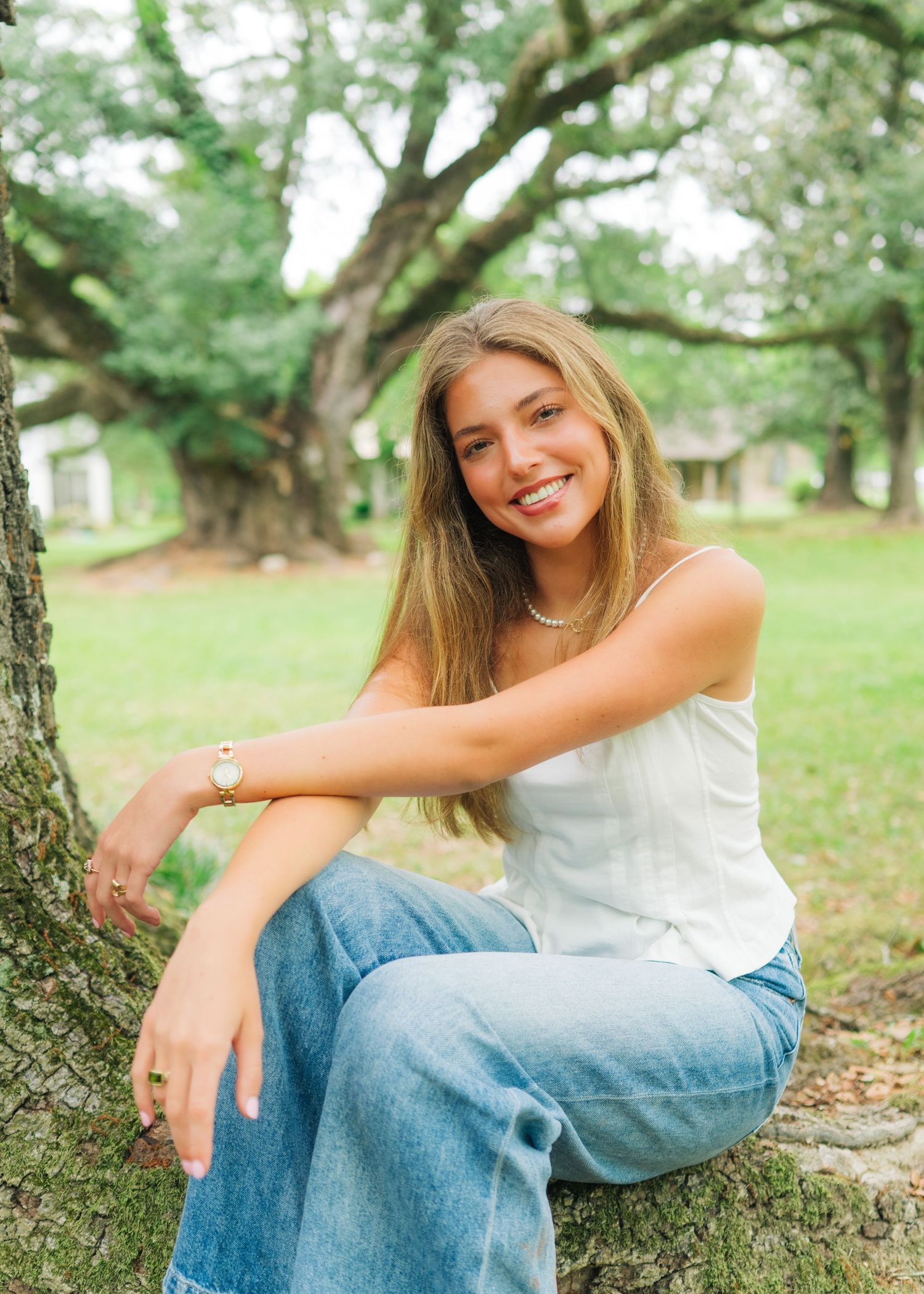 Young woman smiling, sitting against a tree in a park, wearing a white top and blue jeans on a sunny day.