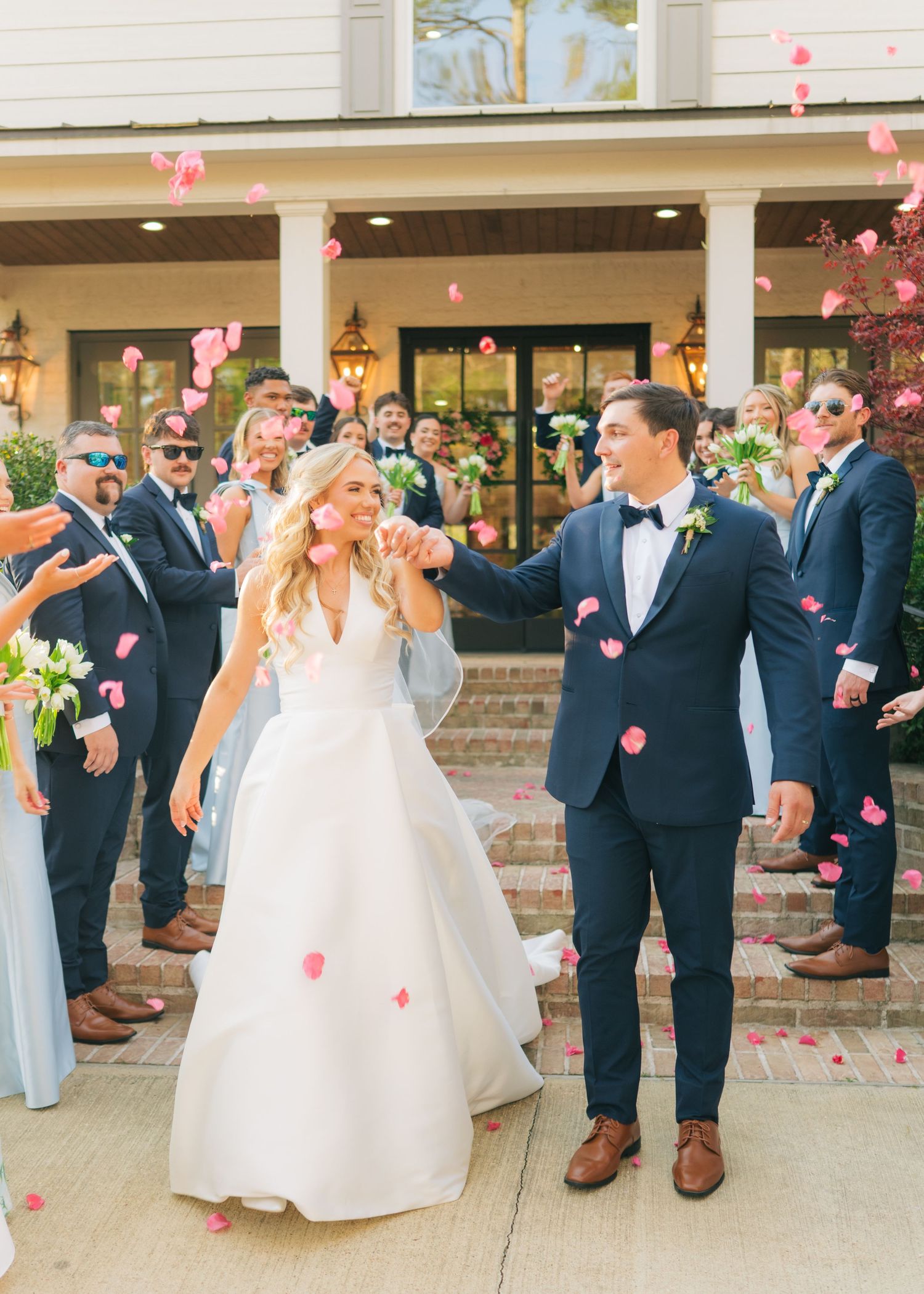 Bride and groom exit ceremony as guests toss pink rose petals outside elegant white estate wedding venue.