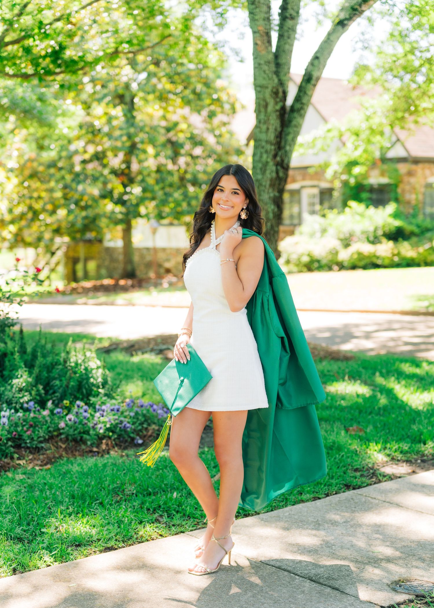 Young woman in white mini dress holding green graduation cap and clutch, posing outdoors in sunny garden setting.