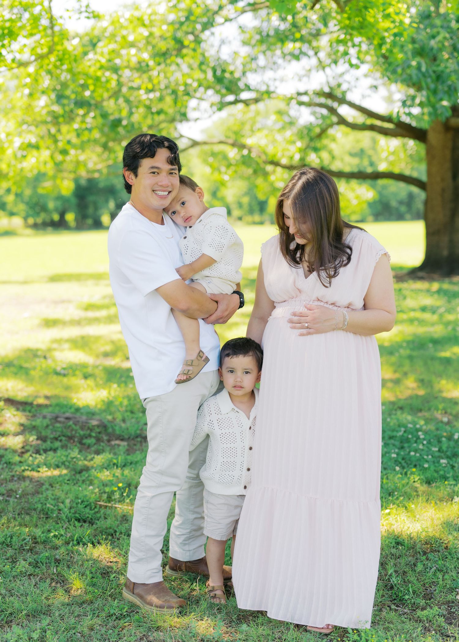 A happy family poses outdoors, with a man holding a baby, a toddler, and a pregnant woman in a pink dress.