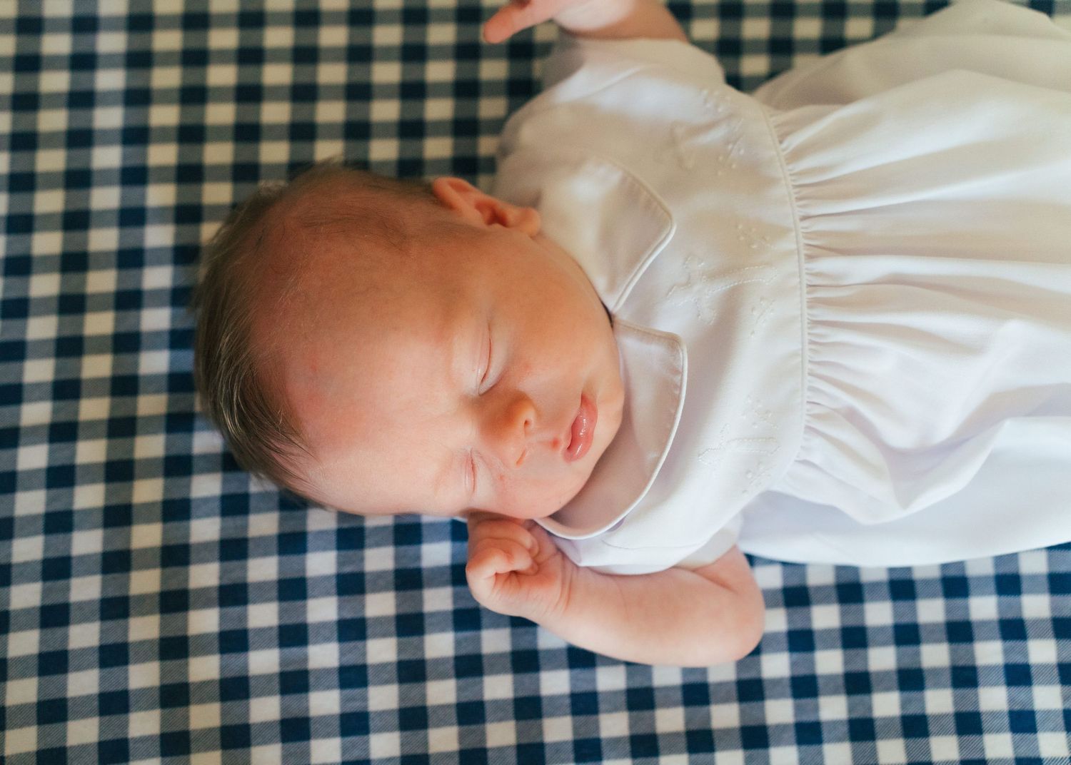 Peaceful newborn baby sleeping in white outfit on blue gingham blanket.
