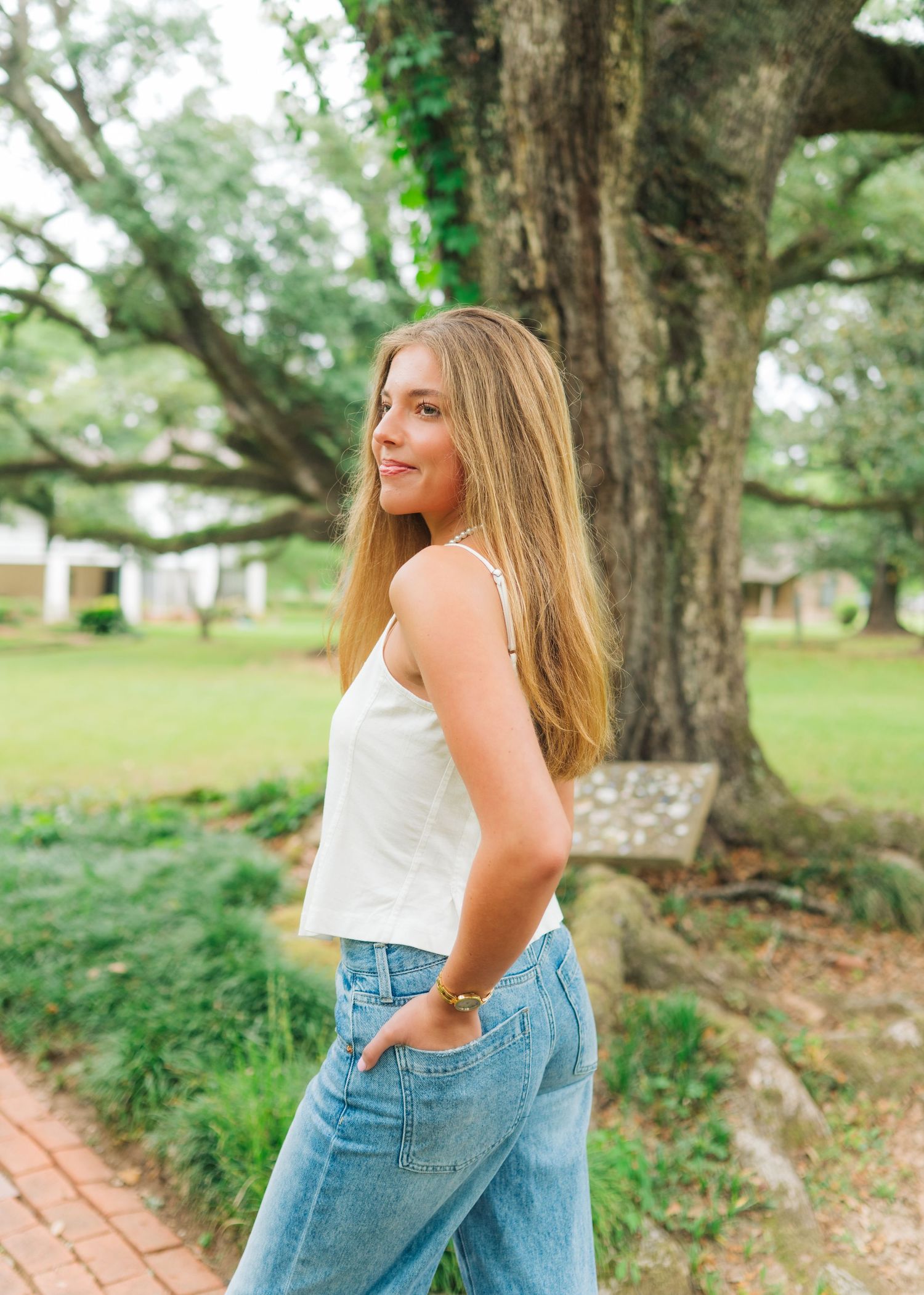 Young woman in white crop top and wide-leg jeans poses outdoors near a large oak tree on a brick path.