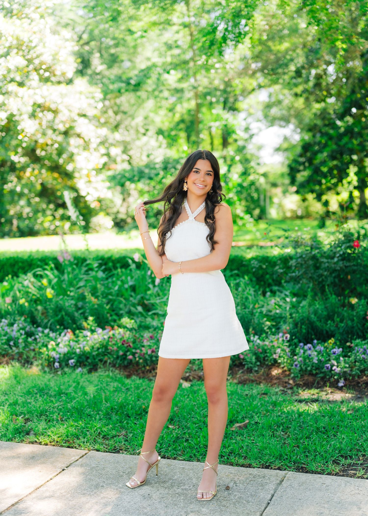 Young woman in white halter dress posing in lush green garden with colorful flowers and stone path.