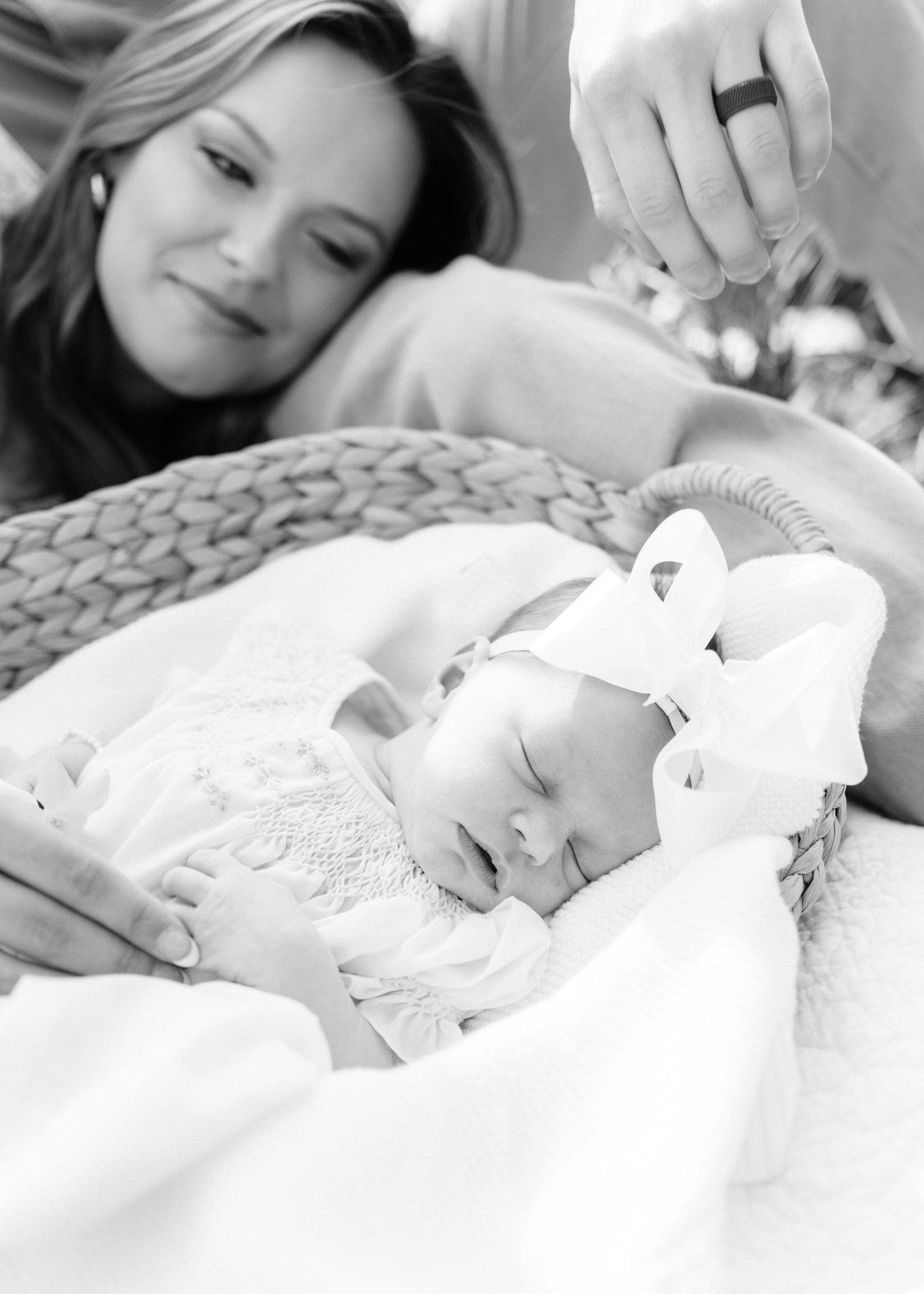 Black and white photo of parents lovingly gazing at their sleeping newborn baby girl in a wicker basket.