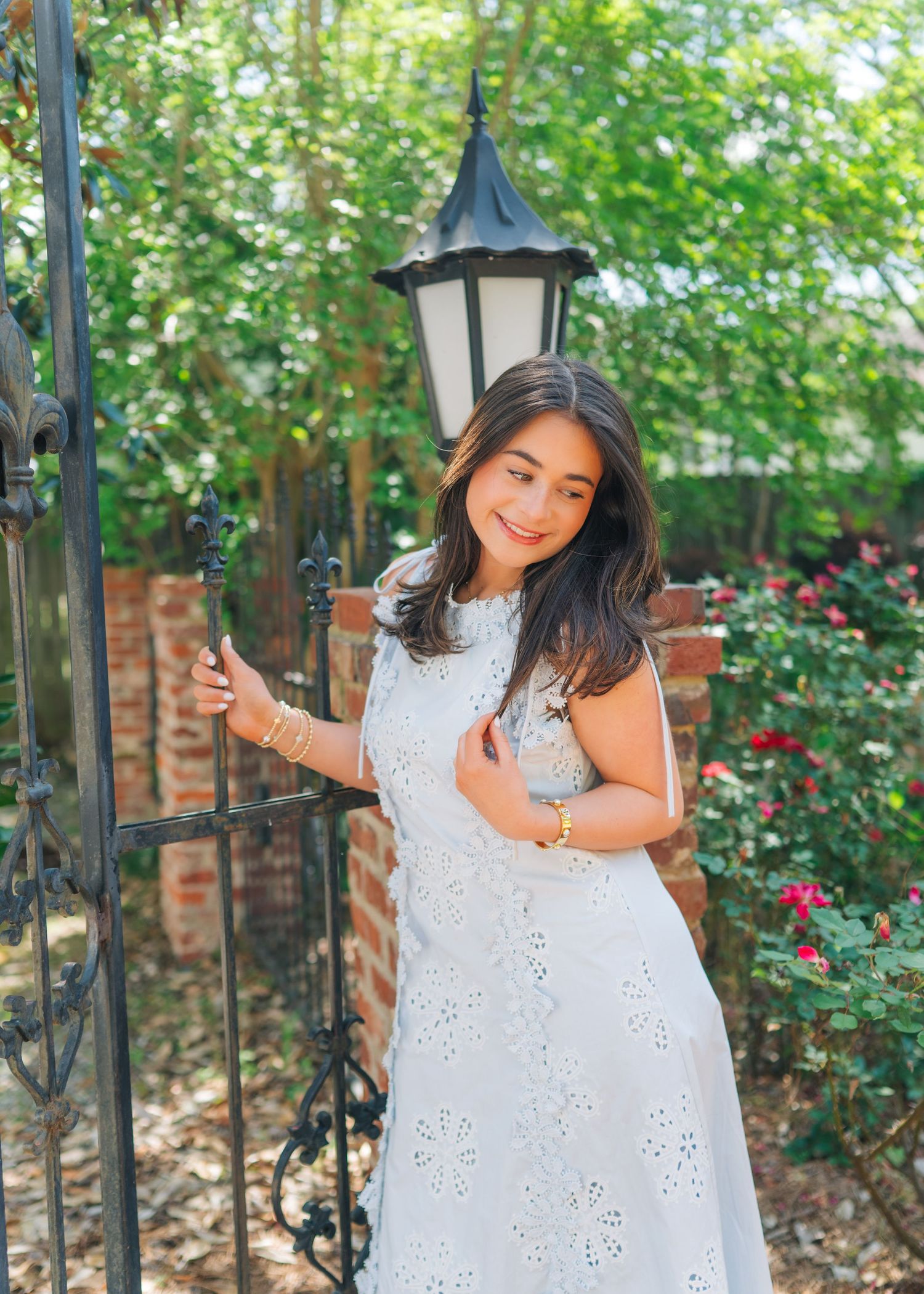 Smiling young woman in white eyelet dress posing by a wrought iron gate with a garden lantern.