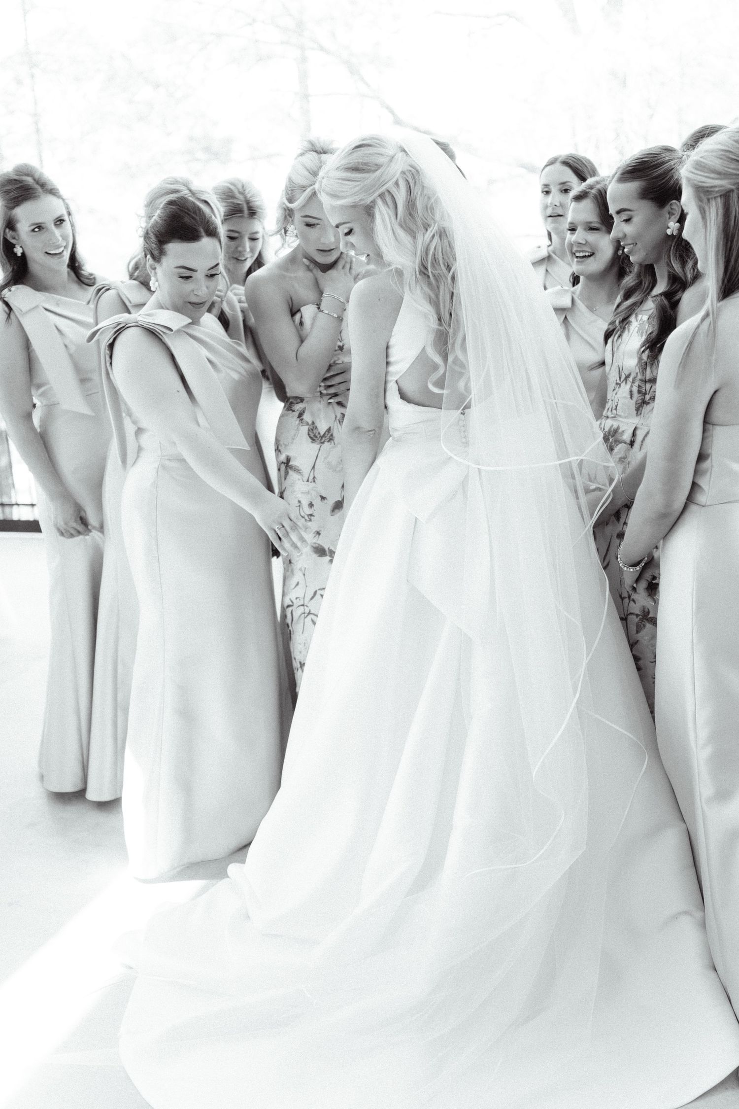 Bride in white gown and veil surrounded by smiling bridesmaids in black and white photo.