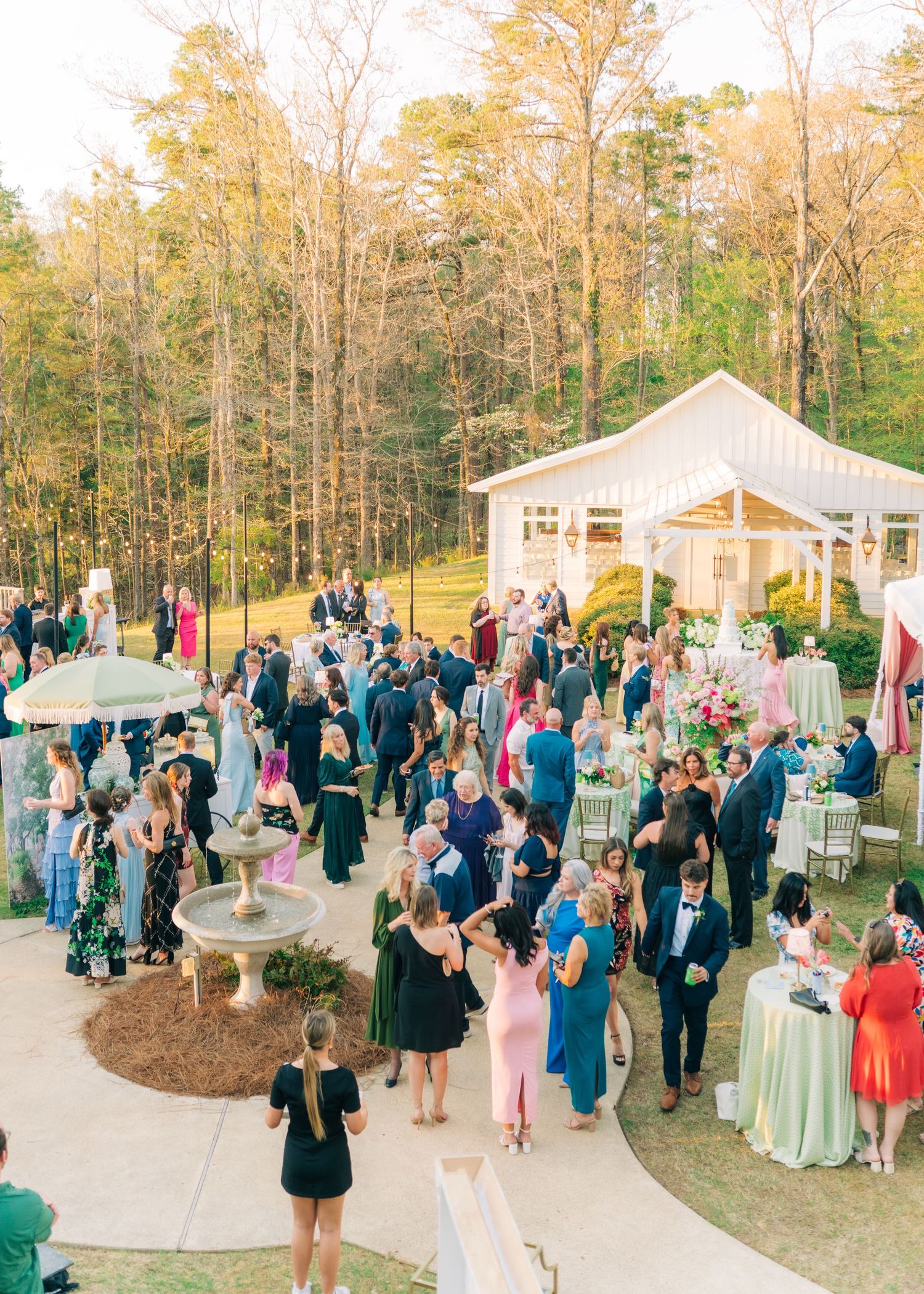 Elegant outdoor wedding reception with guests mingling on a patio surrounded by tall trees and a white tent venue.